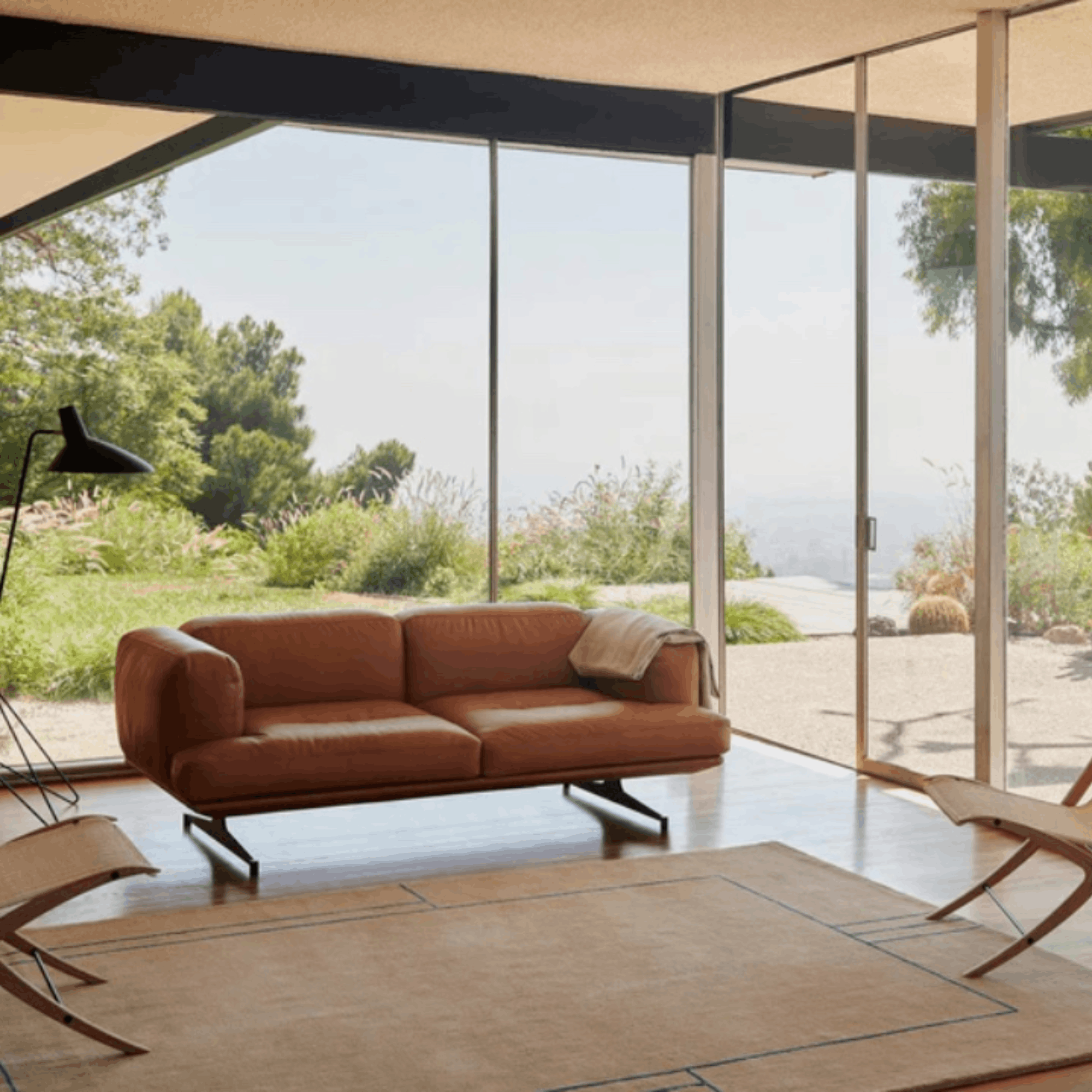A mid-century modern two-seater sofa in warm cognac brown leather with black metal legs, styled in a minimalist, sunlit room with large glass walls overlooking a green landscape.