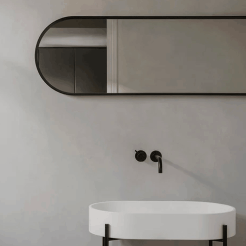 The Audo Copenhagen Norm Mirror hanging on a grey wall above a white marble sink with black taps in a modern bathroom