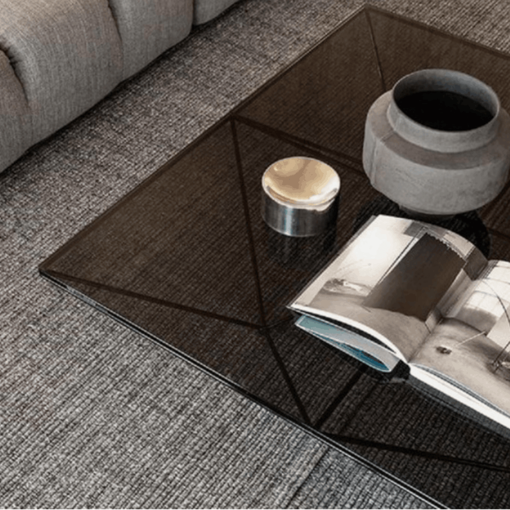 A modern square black glass coffee table with a textured geometric pattern, styled in a living room setting with an open magazine, a small metallic tin, and a gray ceramic vase on top.