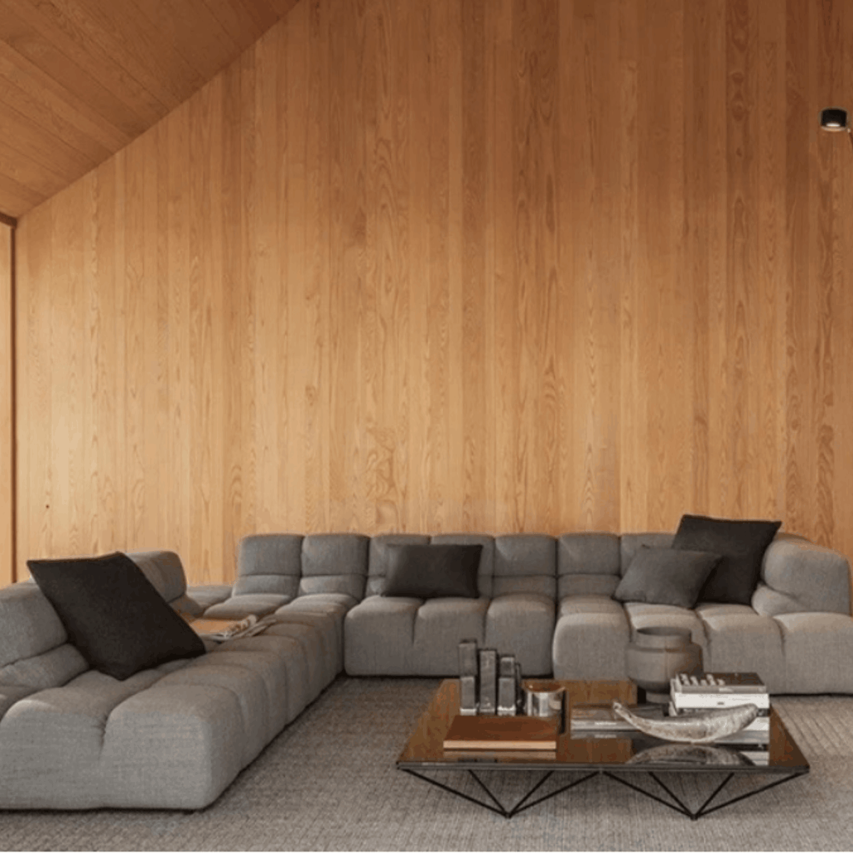 A modern gray modular sectional sofa arranged in an L-shape in a minimalist living room with a light wood-paneled wall and a glass-top coffee table.
