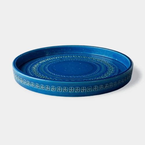 A round ceramic serving tray in a vibrant cobalt blue, featuring a decorative light blue and white geometric pattern along its rim, set against a plain light gray background.