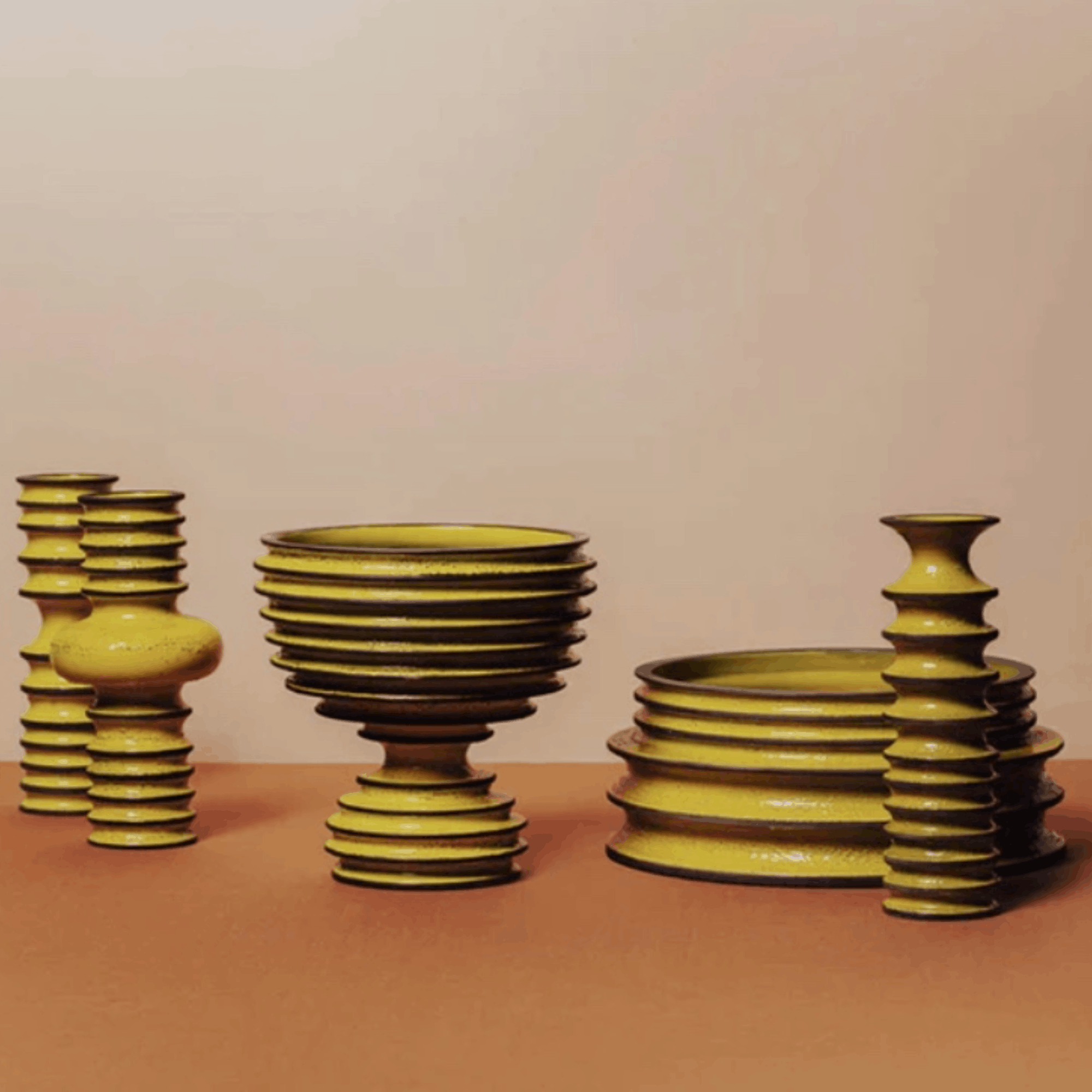 Three groups of stacked ceramic vases and bowls in a glossy olive-yellow glaze, arranged on a terracotta-colored surface against a warm beige background.
