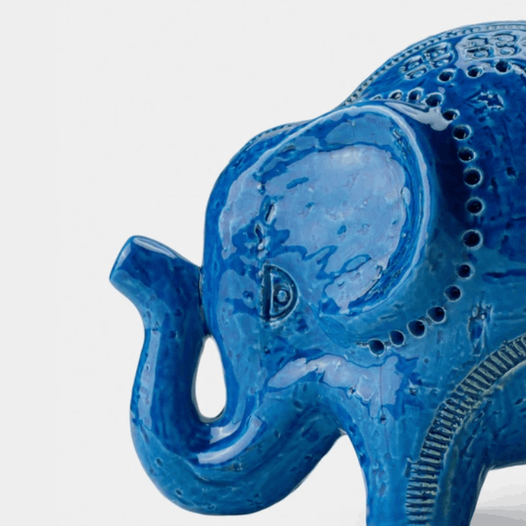 A close-up of a glossy blue elephant figurine with textured details and decorative patterns, shown against a plain light gray background.