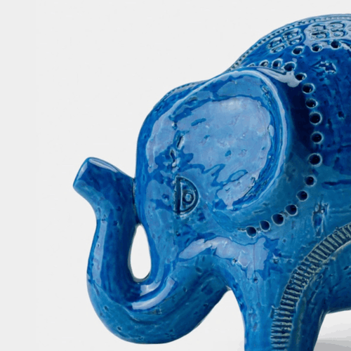 A close-up of a glossy blue elephant figurine with textured details and decorative patterns, shown against a plain light gray background.