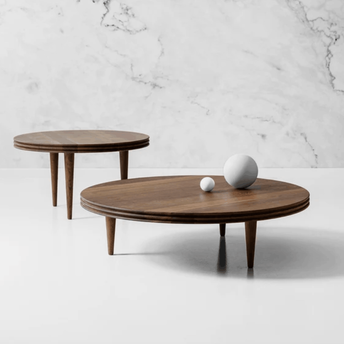 Two round dark wood coffee tables with tapered legs, styled with white decorative spheres on a white floor against a white marble wall.