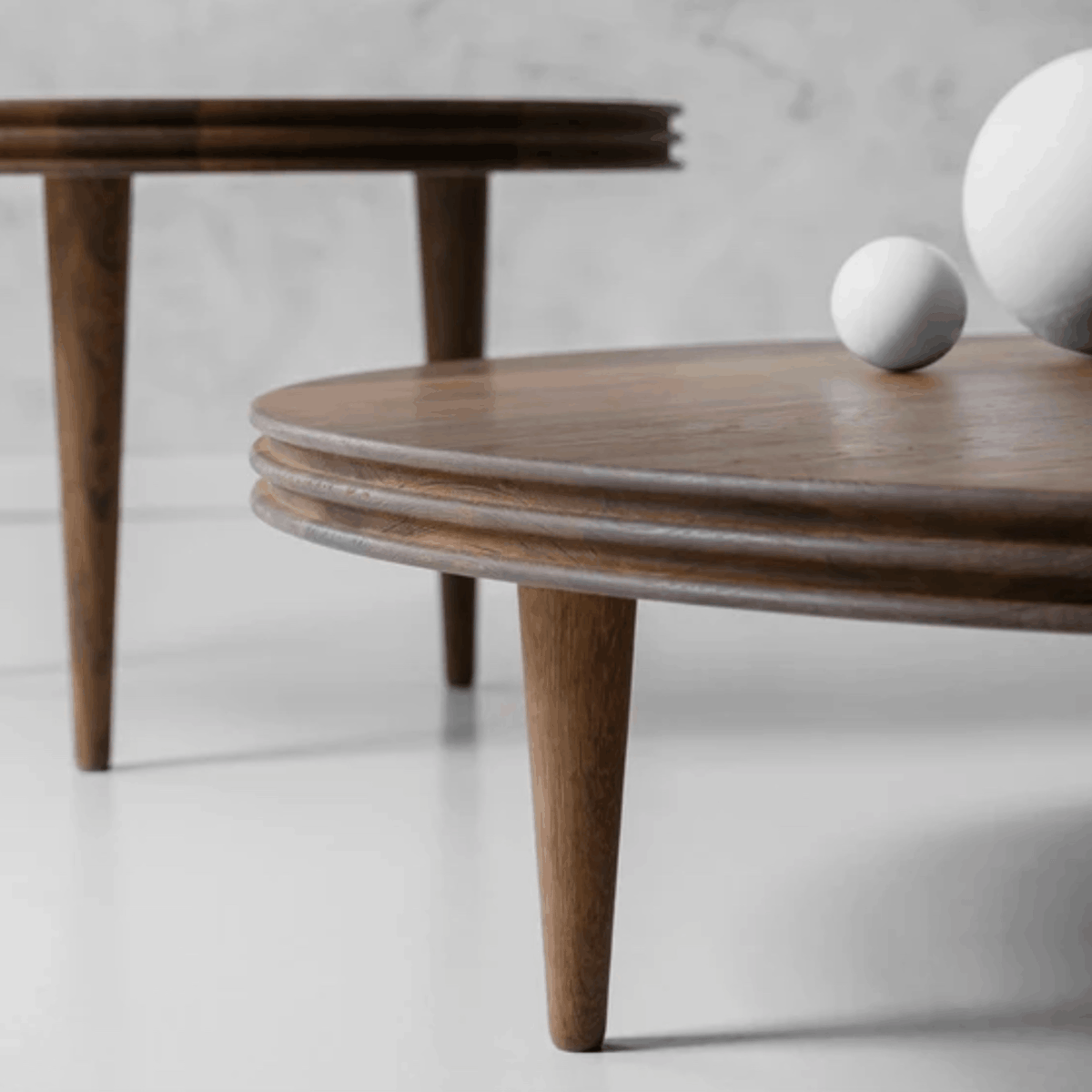 A close-up of a round wooden coffee table with layered edges and tapered legs, shown in a minimalist interior setting with white decorative spheres on top.