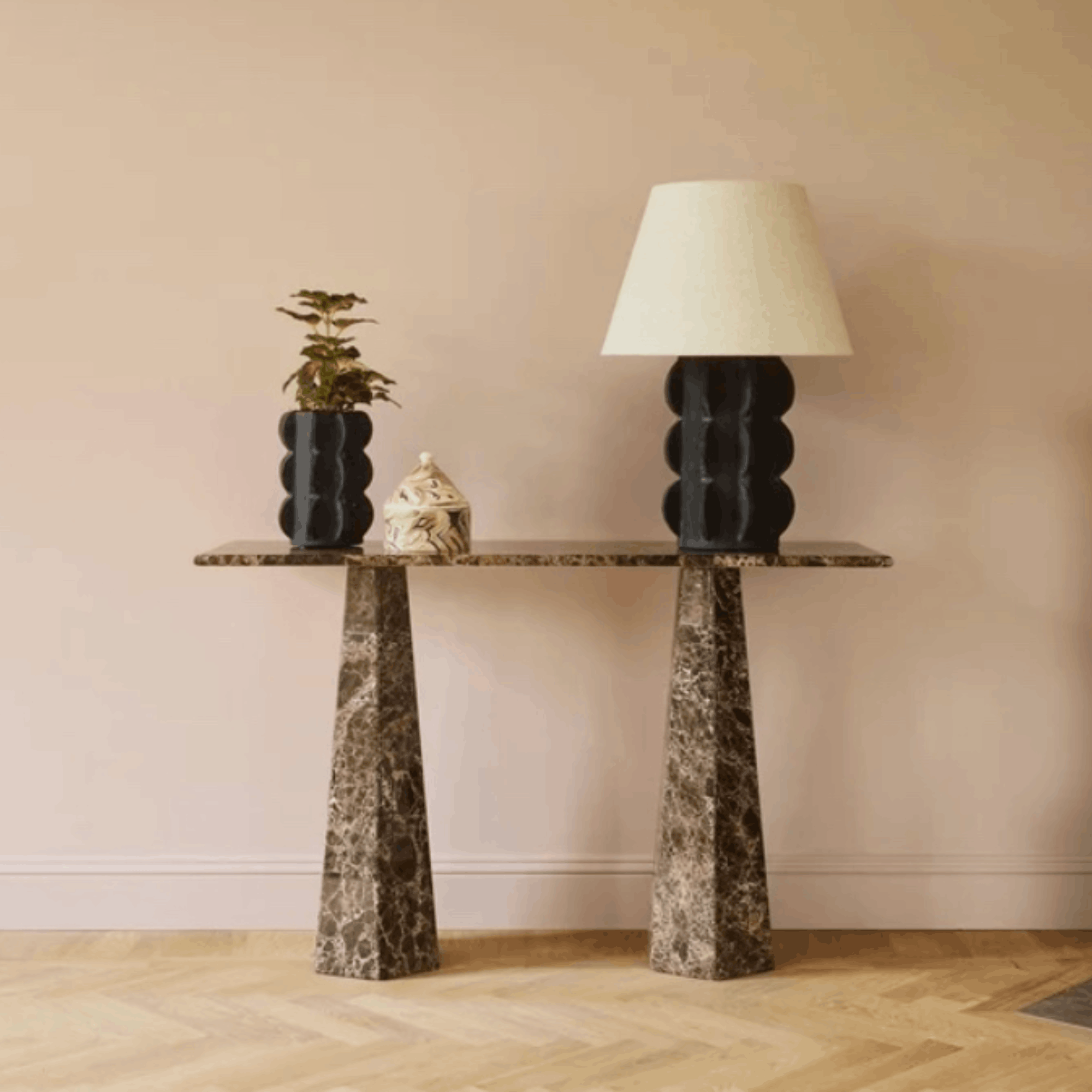 A modern console table with a dark marble top and two tapered stone legs, styled with a black sculptural table lamp, a small potted plant, and a decorative box against a beige wall and light wood floor.