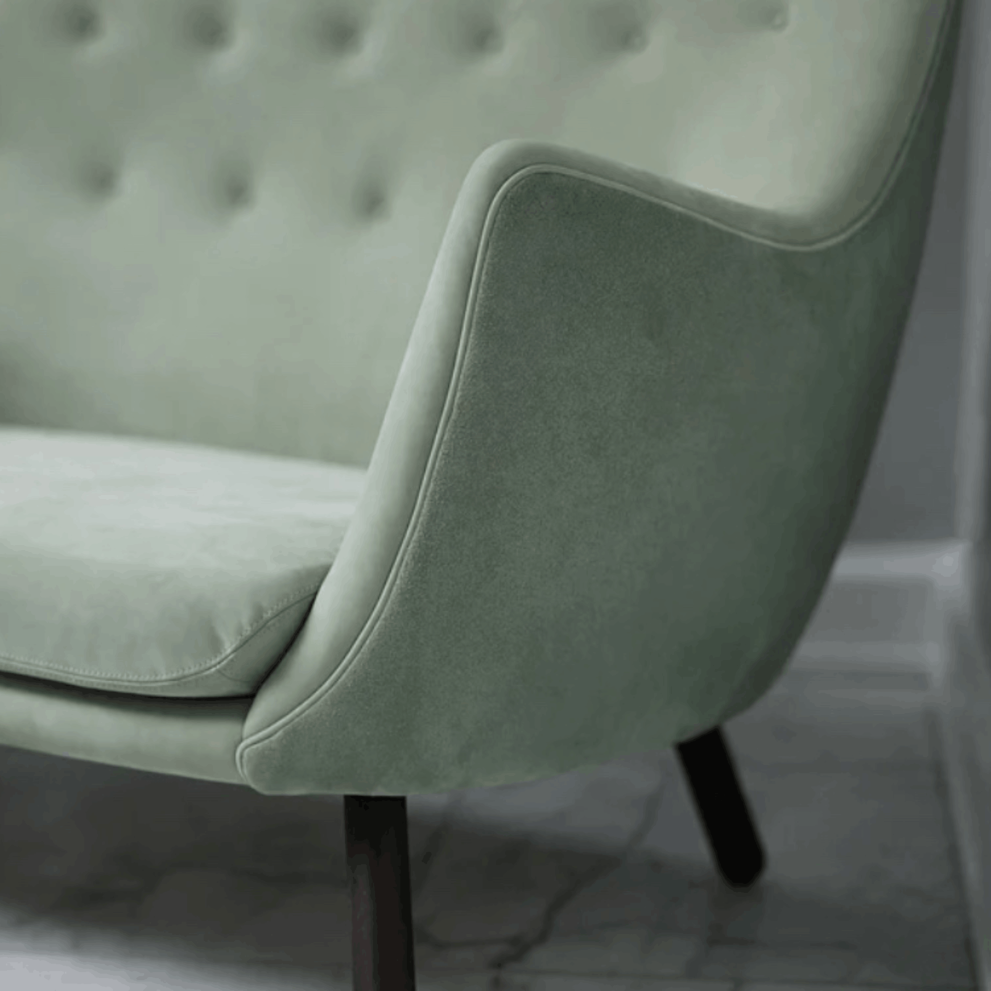 A close-up of a light sage green velvet armchair with a tufted backrest and dark wooden legs, set against a neutral gray wall and marble floor.