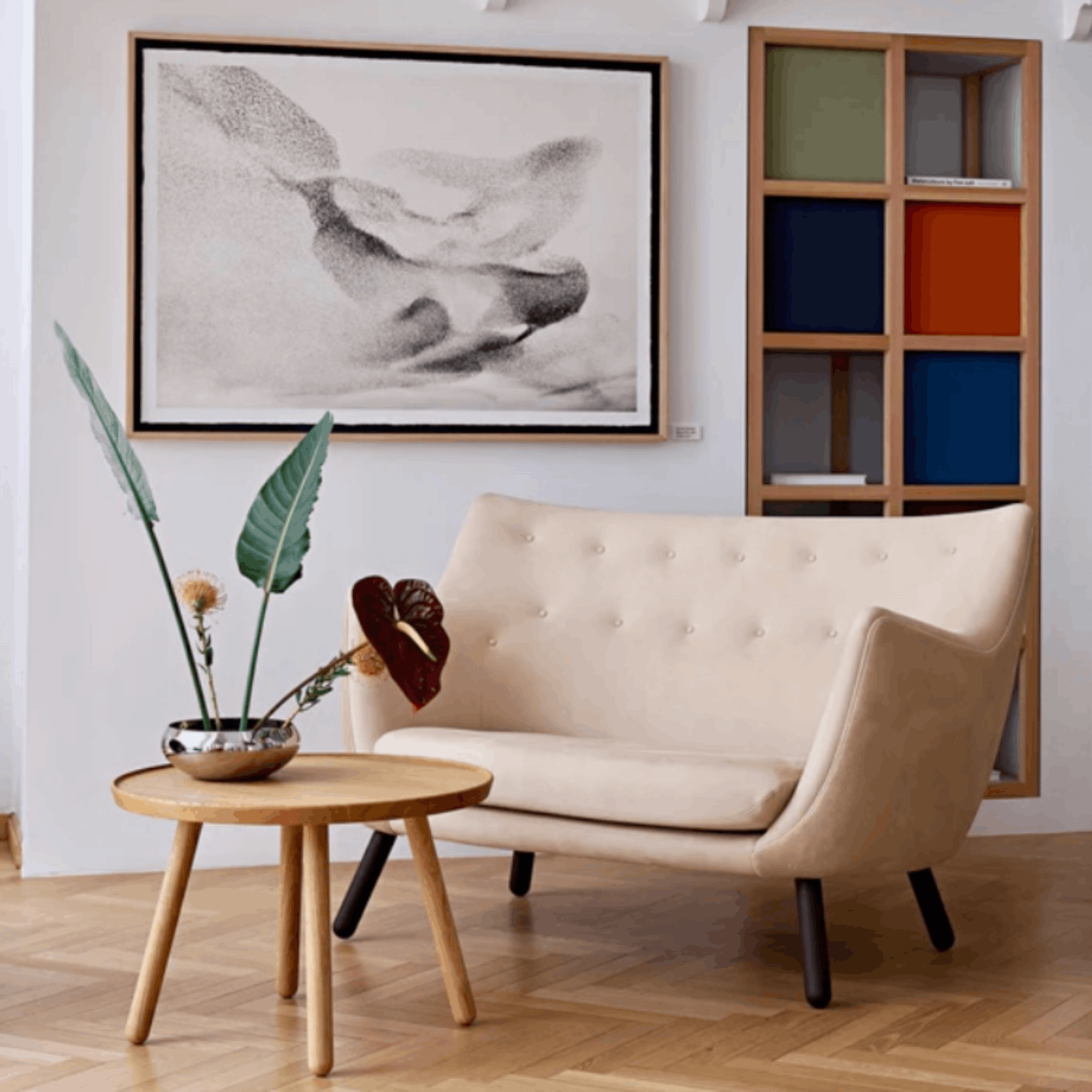A cream tufted loveseat sofa in a modern living room setting, styled with a round wooden side table holding a vase with green leaves, a large black and white framed abstract print on the wall, and a wooden shelving unit with colorful compartments.