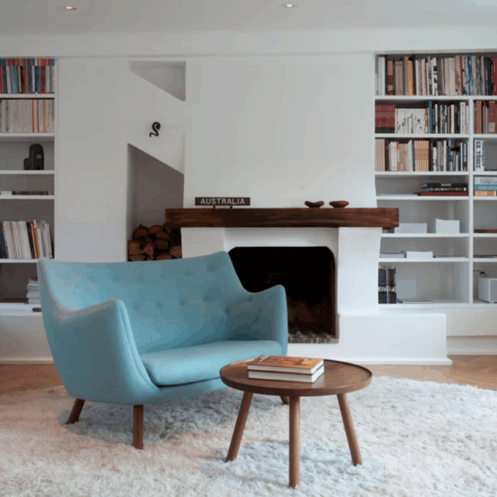 A mid-century modern light blue velvet armchair in a bright, white-walled living room with a fireplace, bookshelves, and a small round wooden coffee table.
