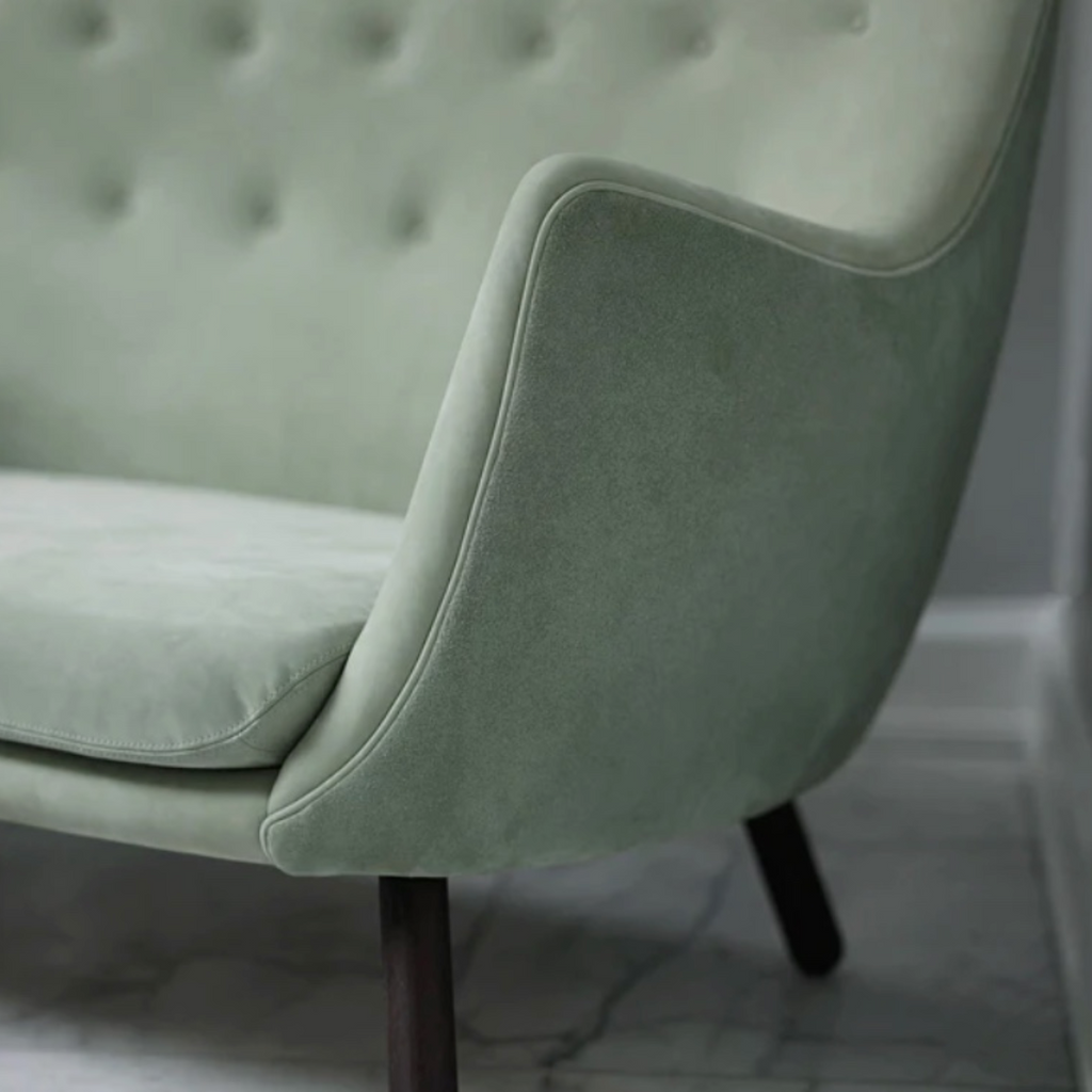 A close-up of a light sage green velvet armchair with a tufted backrest and dark wooden legs, set against a neutral gray wall and marble floor.