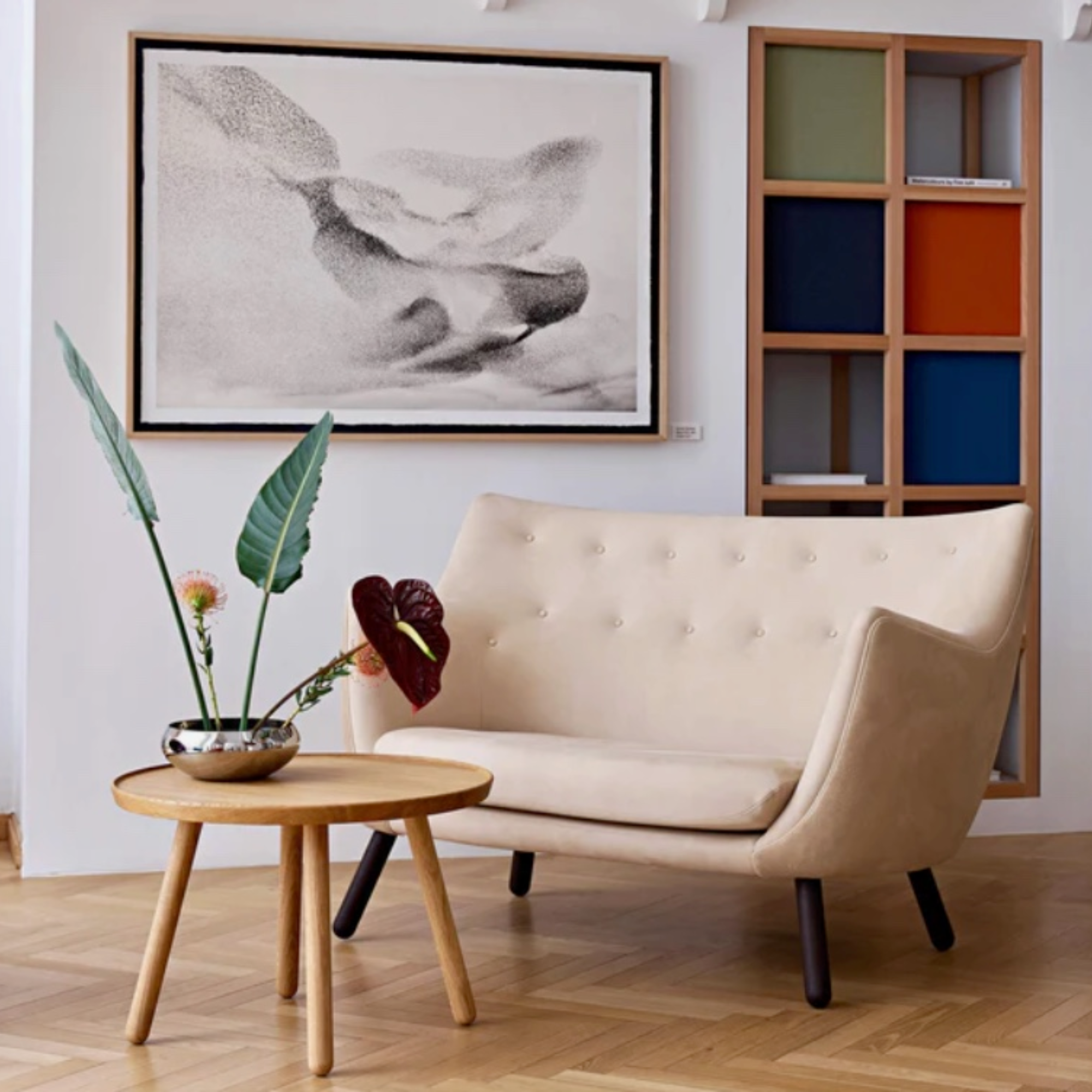 A cream tufted loveseat sofa in a modern living room setting, styled with a round wooden side table holding a vase with tropical leaves, a large black and white framed wall art piece, and a wooden shelving unit with colorful panels.