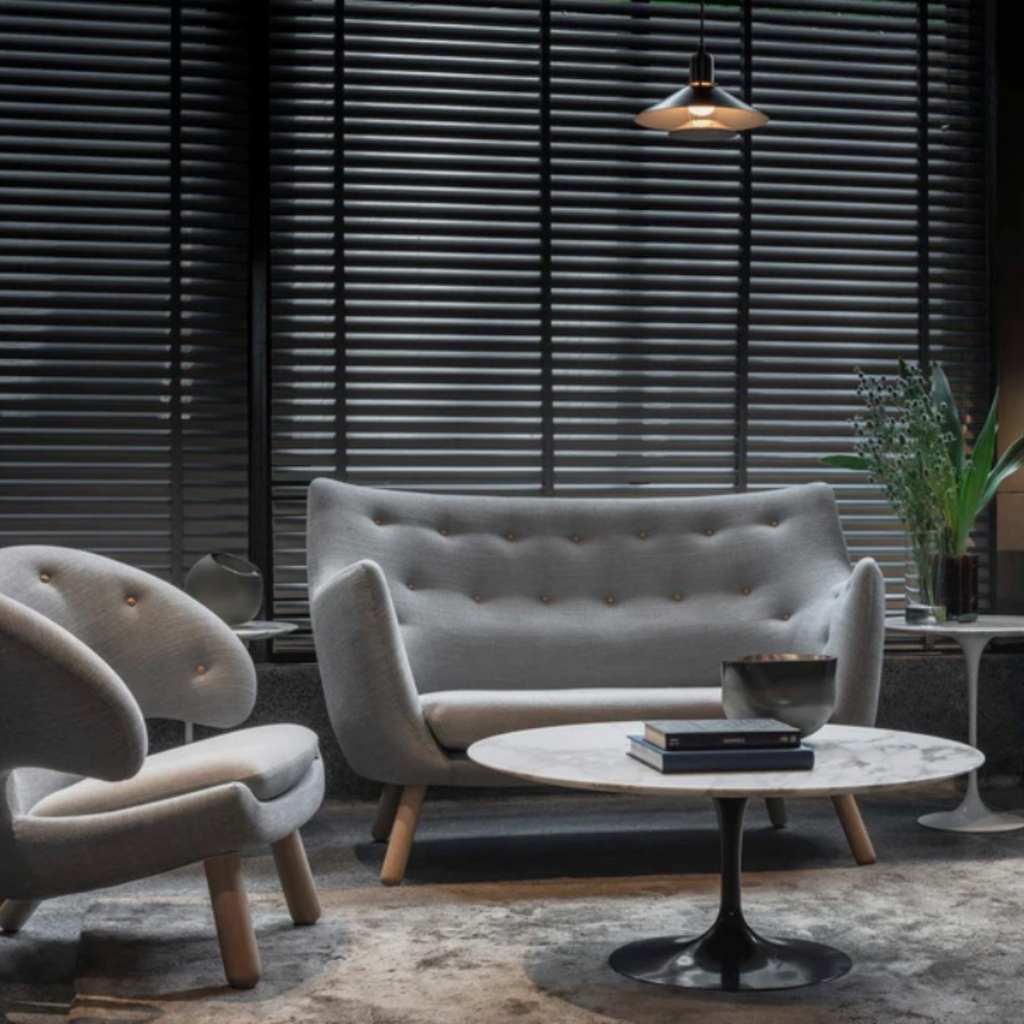 A modern living room scene featuring a light gray tufted loveseat, a matching curved armchair, a round marble-top coffee table with a black pedestal base, and dark window blinds in the background.