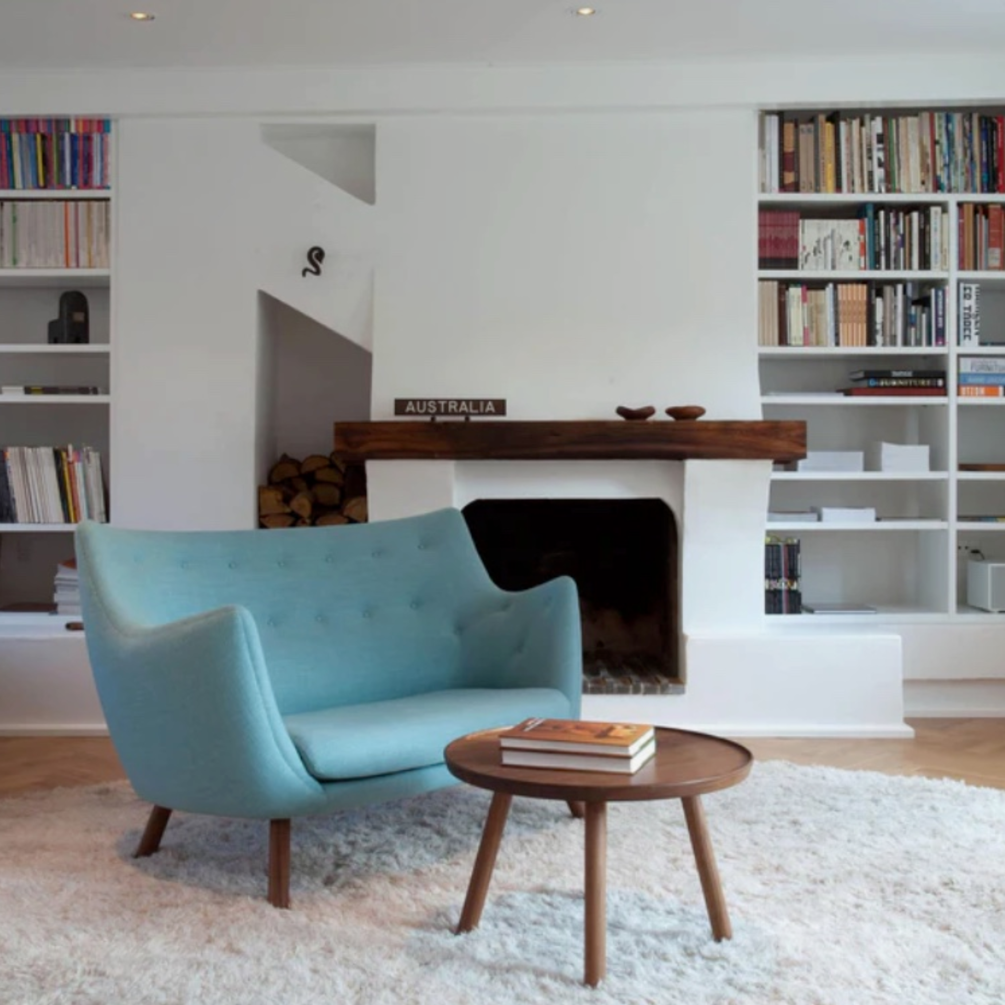 A mid-century modern light blue velvet armchair in a bright, white-walled living room with a fireplace, bookshelves, and a small round wooden coffee table.