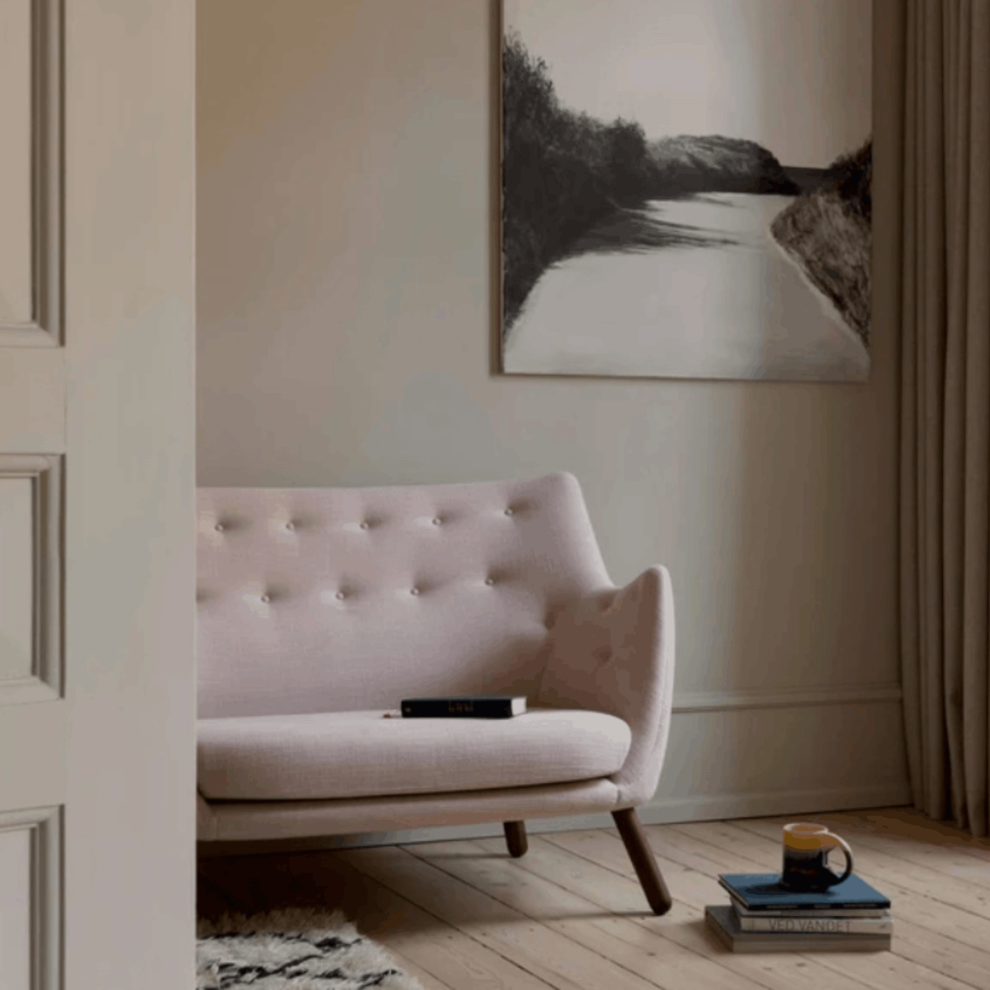 A light blush pink tufted armchair in a styled room setting with a black and white landscape painting on the wall, a mug on books, and a wooden floor.
