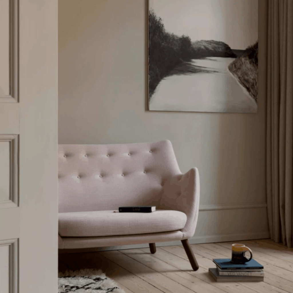 A light blush pink tufted armchair in a styled room setting with a black and white landscape painting on the wall, a mug on books, and a wooden floor.