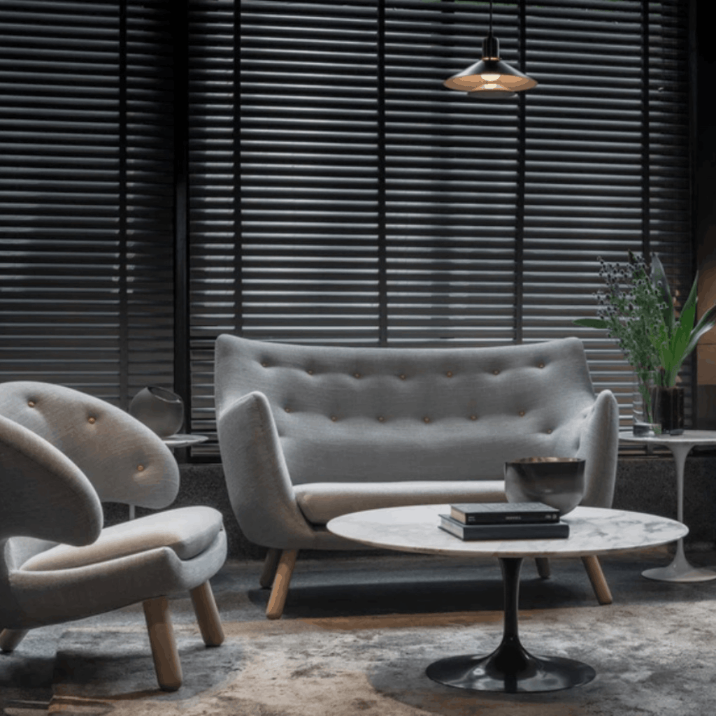A modern living room scene featuring a light gray tufted loveseat, a matching curved armchair, a round marble-top coffee table with a black pedestal base, and dark window blinds in the background.