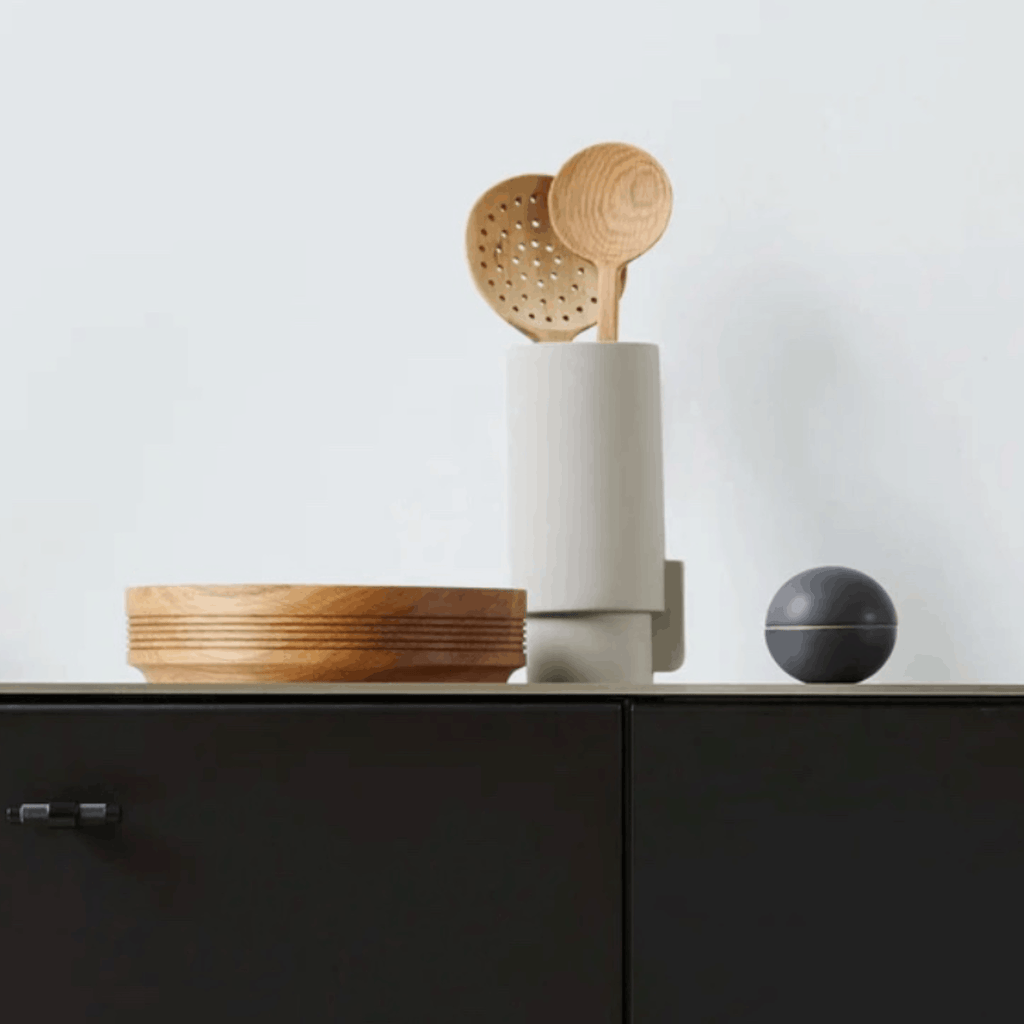 A modern kitchen scene with a white cylindrical vase containing two light wood spoons, placed on a dark countertop beside stacked wooden bowls and a small dark spherical object against a light wall.