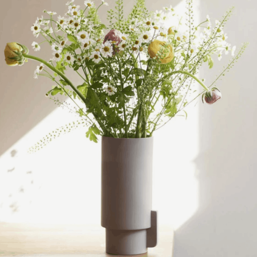 A modern cylindrical gray vase holding a bouquet of white daisies, green foliage, and a few yellow and pink buds, set against a softly lit cream wall.