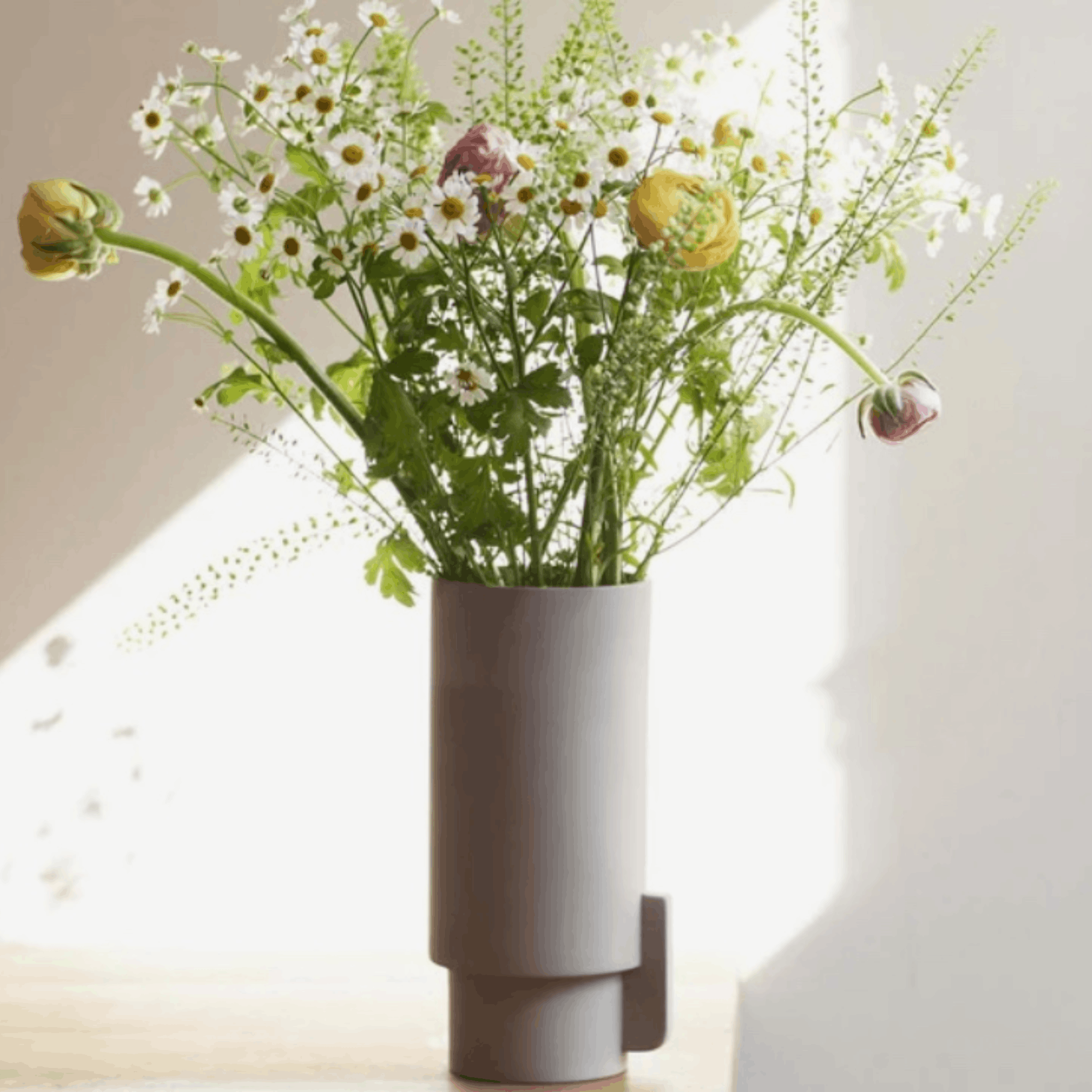 A modern cylindrical gray vase holding a bouquet of white daisies, green foliage, and a few yellow and pink buds, set against a softly lit cream wall.