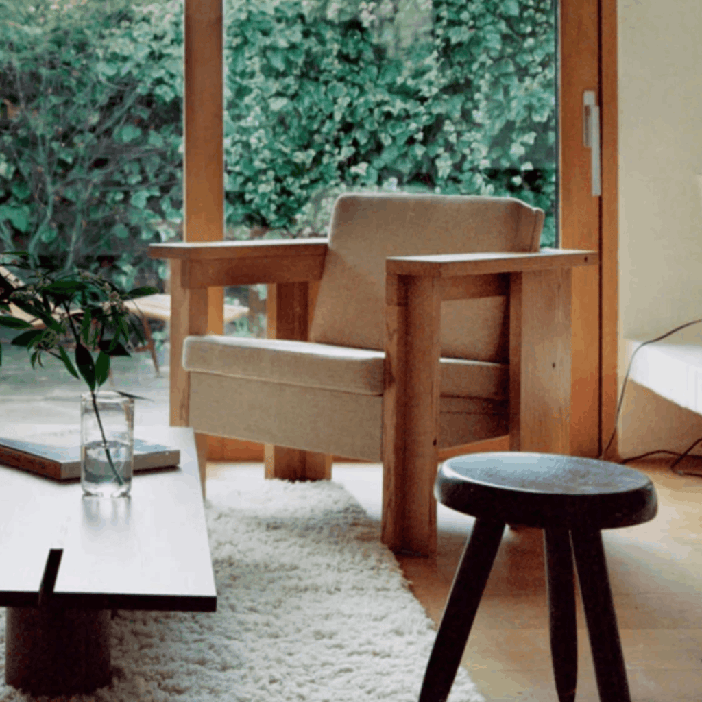Modern living room with wooden armchair, side table, and stool near a glass door with greenery outside.