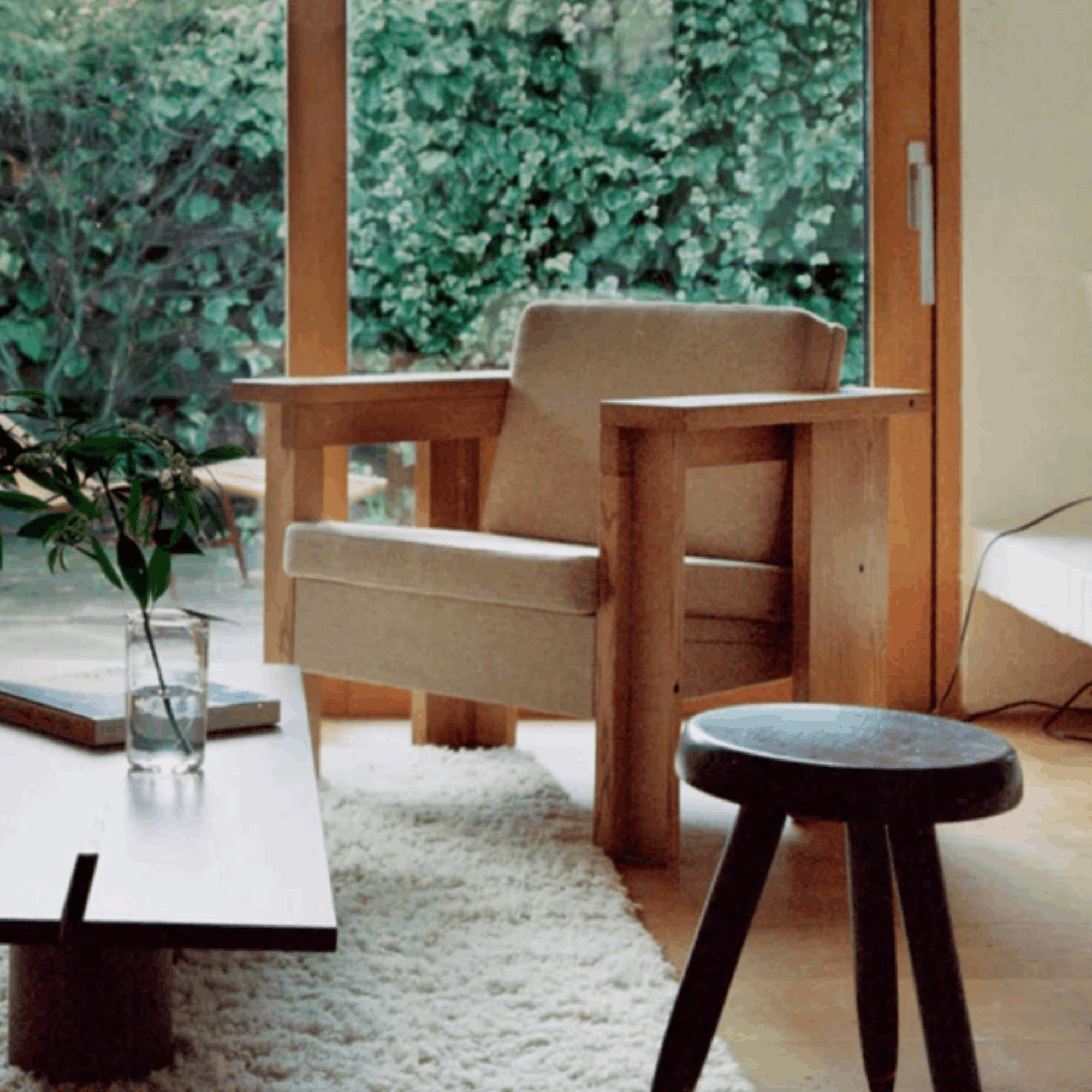 Modern living room with wooden armchair, side table, and stool near a glass door with greenery outside.