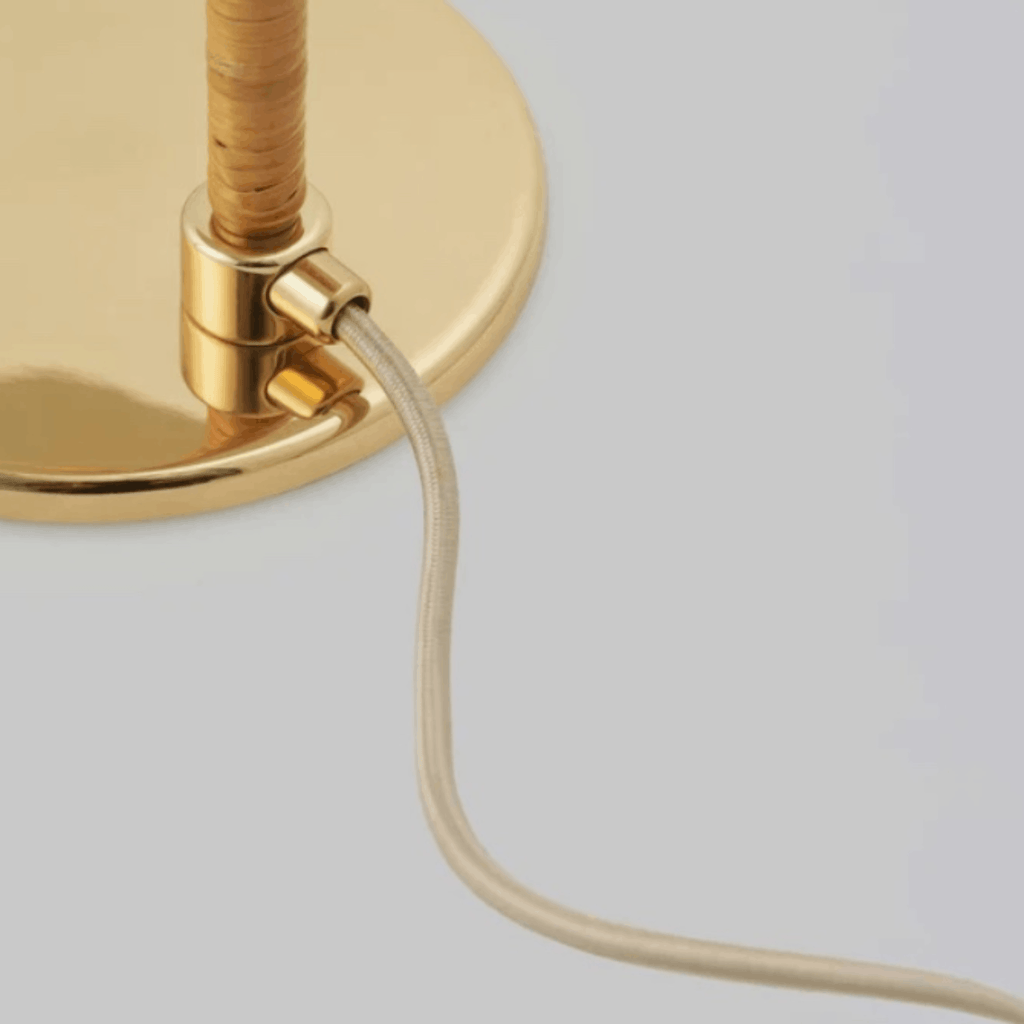 Close-up of a brass table lamp base with a beige fabric cord emerging from the side, set against a solid light gray background.