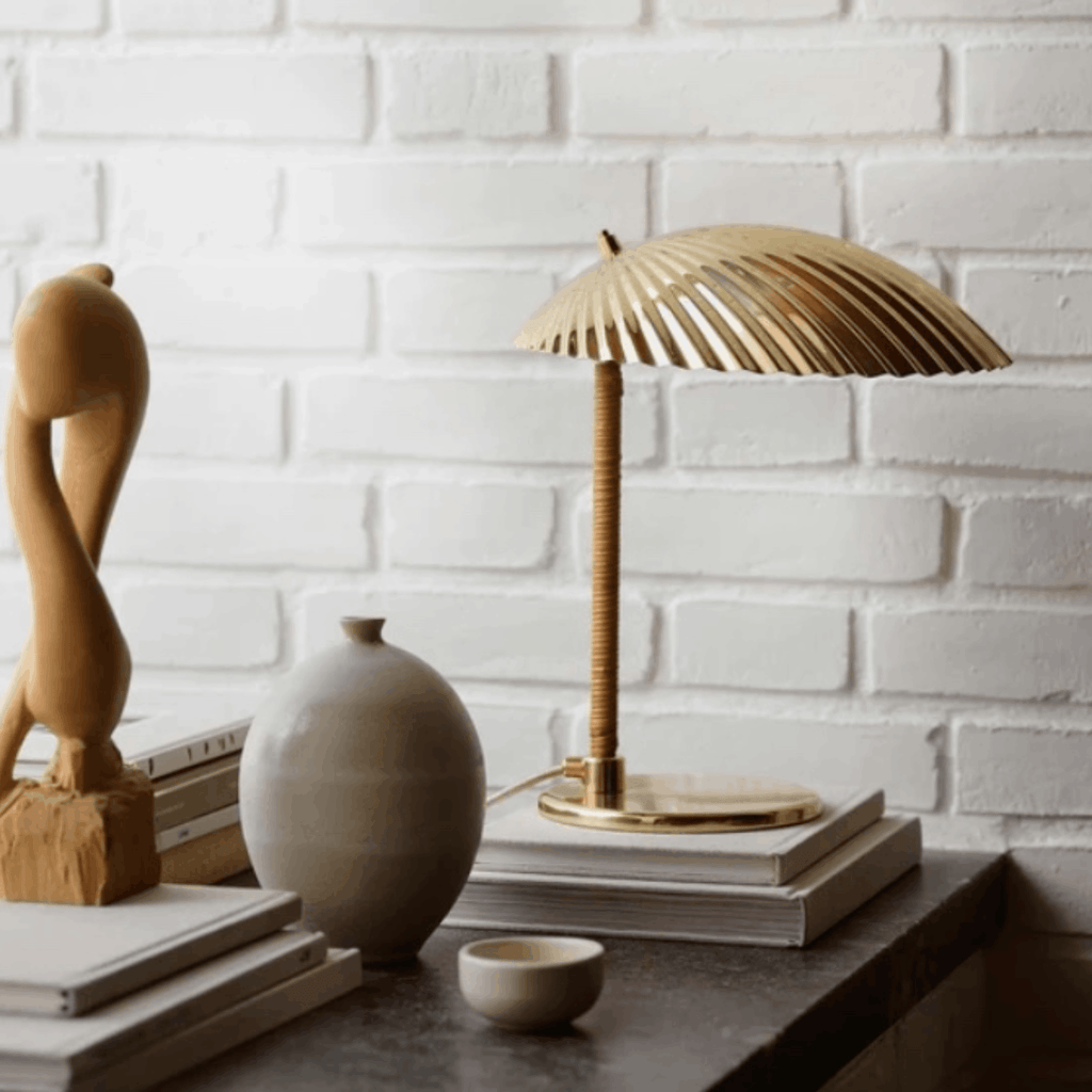 A modern table lamp with a gold-toned slatted dome shade and a textured gold stem, styled on a dark tabletop with books, a ceramic vase, and a small bowl against a white brick wall.