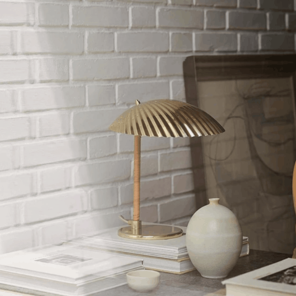 A modern table lamp with a ribbed metallic gold dome shade and a light wood stem, styled on a tabletop next to a ceramic vase and books against a white brick wall.