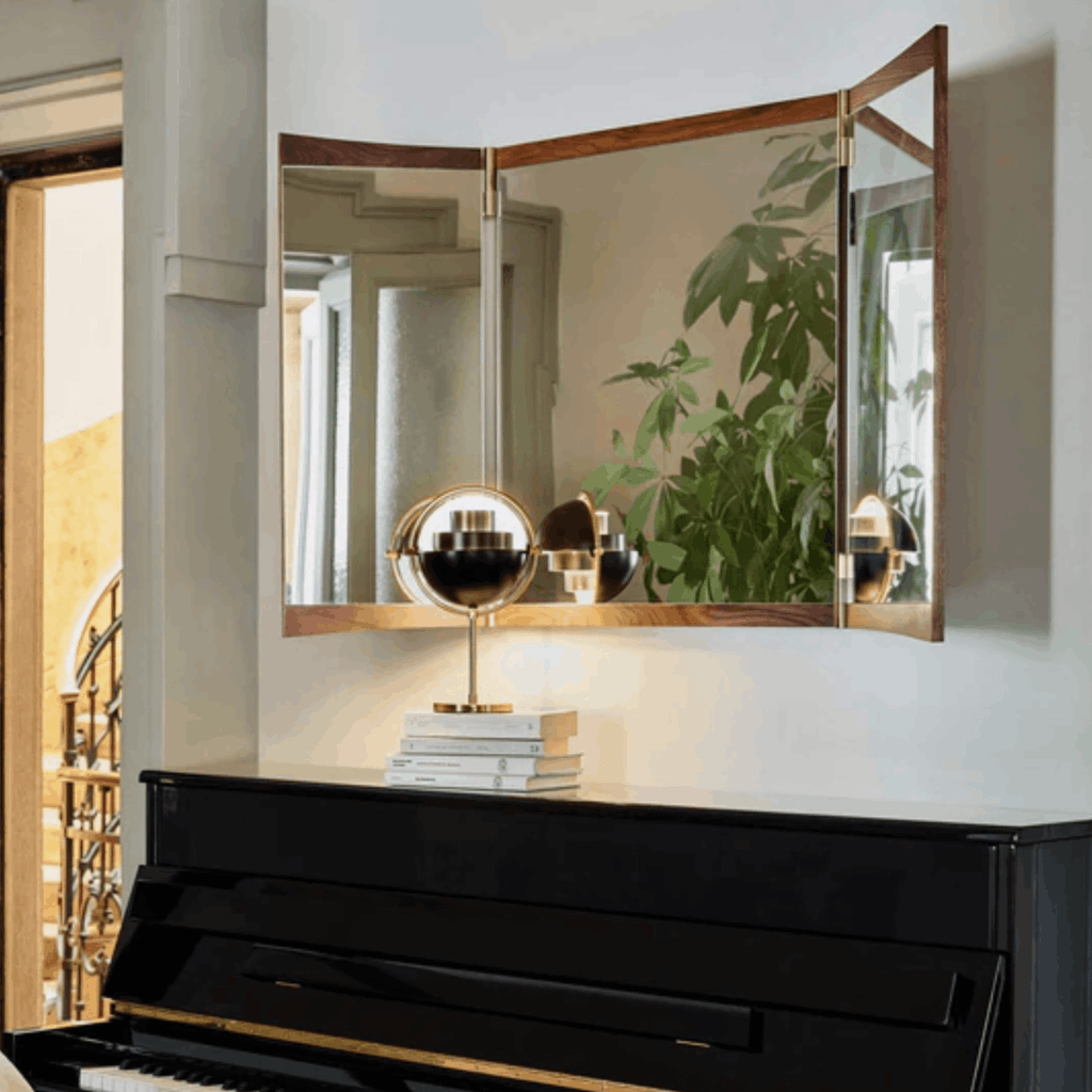 A three-panel folding wall mirror with a warm wood frame hangs above a black piano in a styled interior. The mirror reflects green plants and a gold table lamp, and the room has light neutral walls.