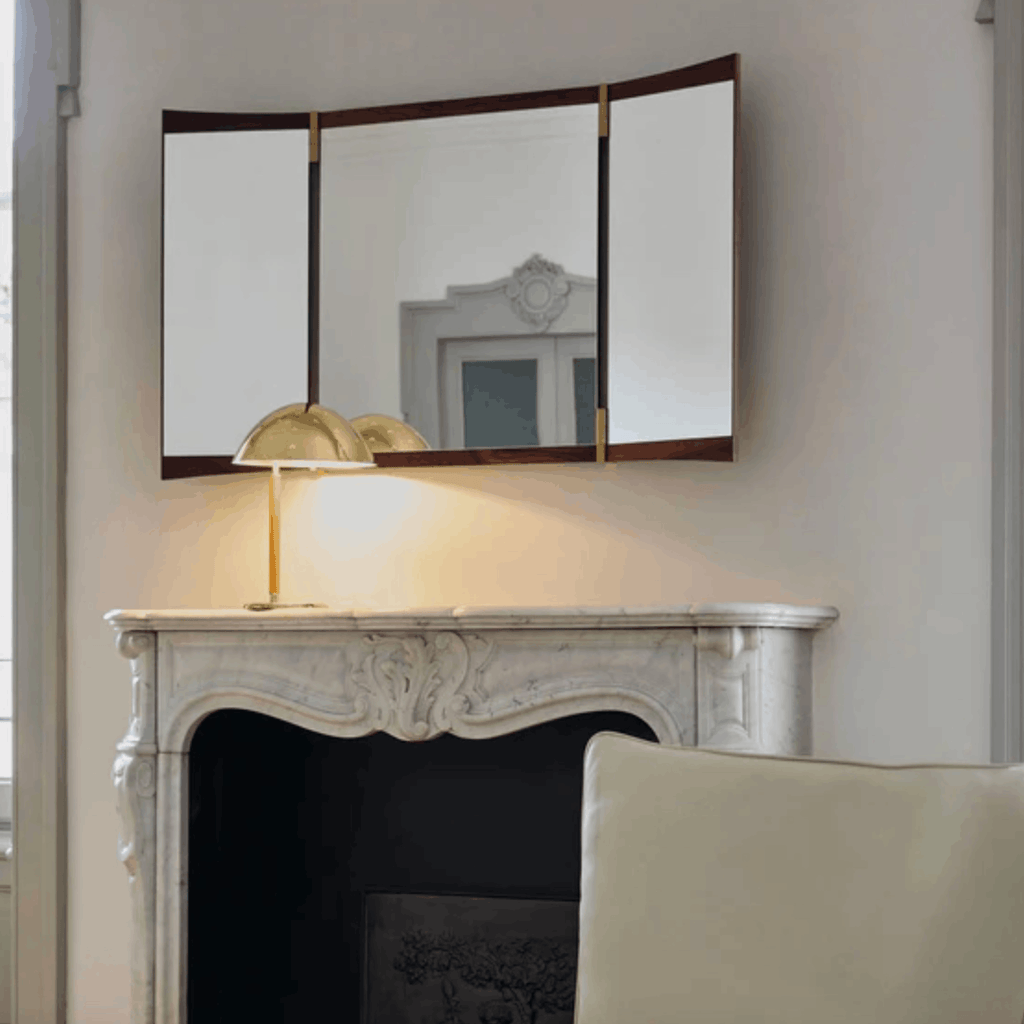 A three-panel folding wall mirror with a dark wood frame and gold-tone hinges, mounted above a white marble fireplace mantel. A gold table lamp sits on the mantel, and a cream-colored armchair is partially visible in the foreground.