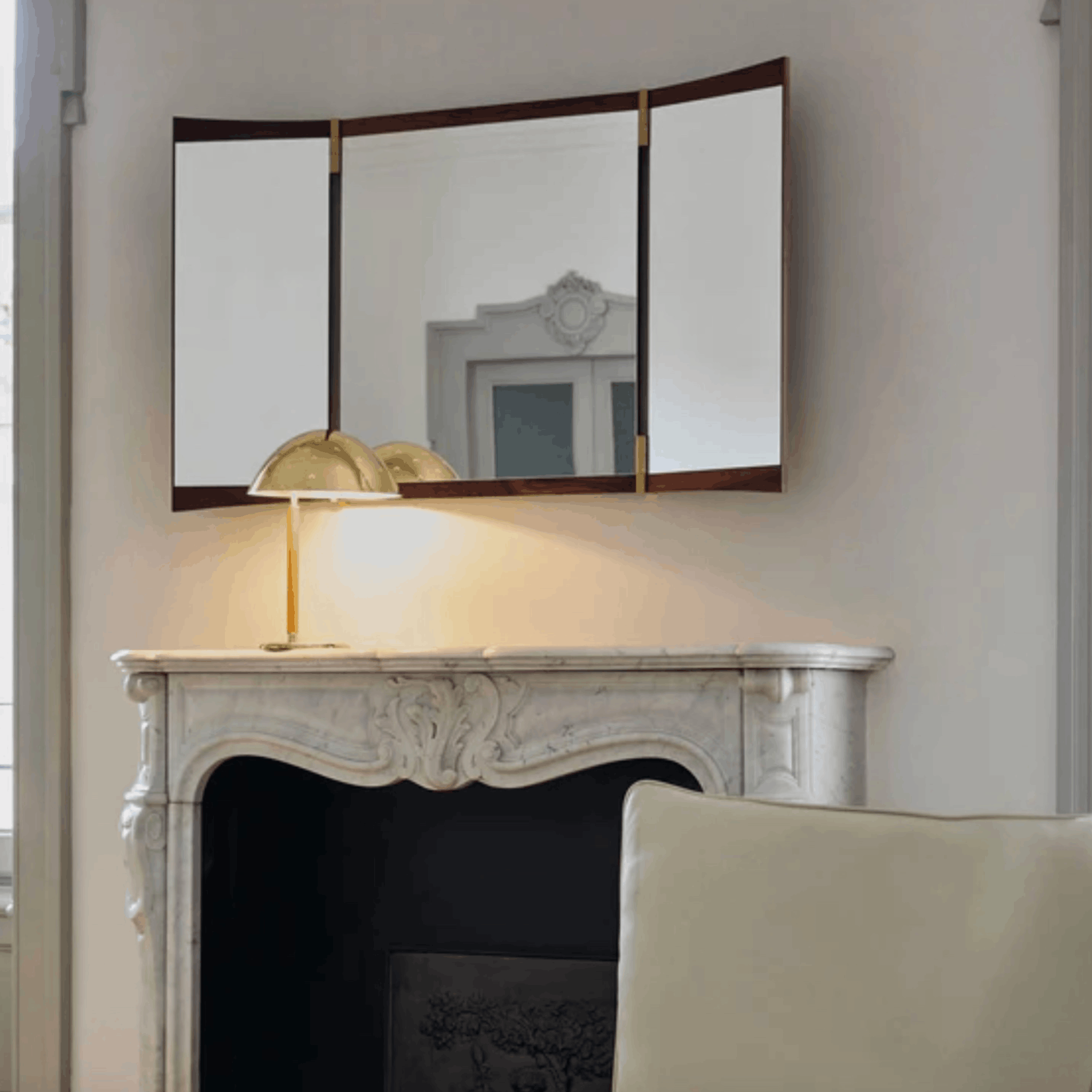 A three-panel folding wall mirror with a dark wood frame and gold-tone hinges, mounted above a white marble fireplace mantel. A gold table lamp sits on the mantel, and a cream-colored armchair is partially visible in the foreground.