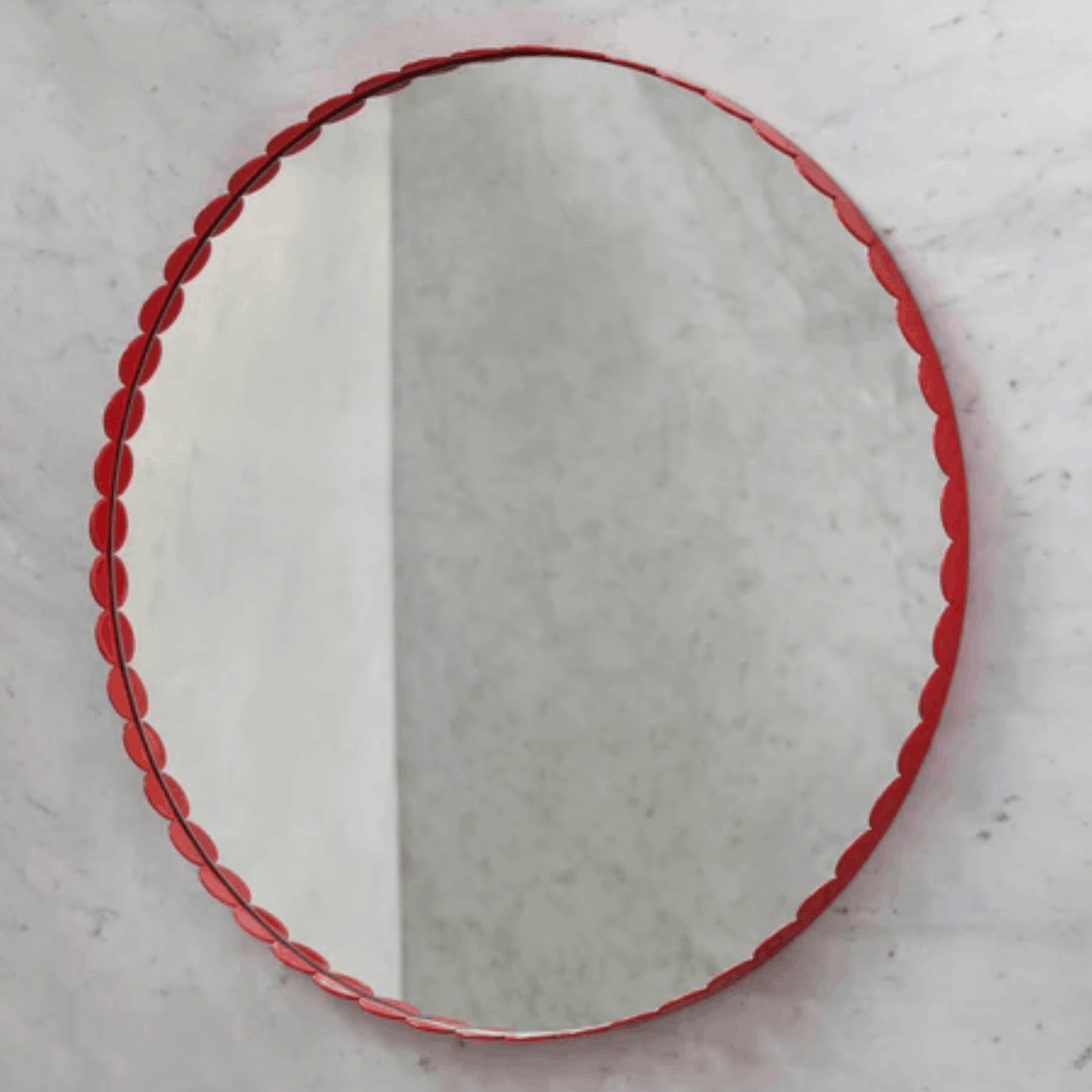 An oval wall mirror with a decorative red scalloped frame, shown against a light gray textured wall.