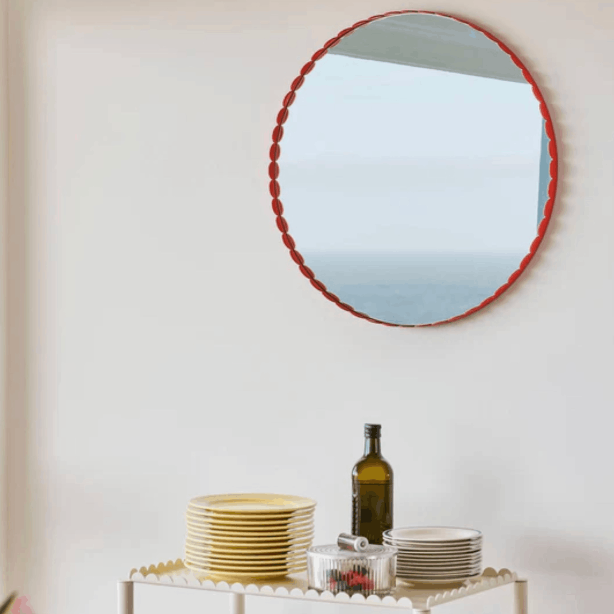 A round wall mirror with a scalloped red frame hangs on a white wall above a white table styled with plates, a glass bottle, and a small bowl.