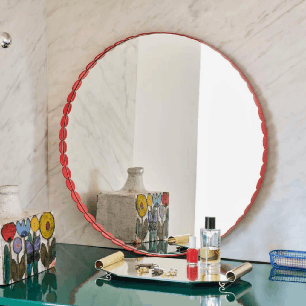 A round wall mirror with a scalloped coral-red rim, leaning on a green glass tabletop against a light marble wall. The mirror reflects a bright white wall and a decorative vase, while the tabletop below holds small bottles, jewelry, and a tray.