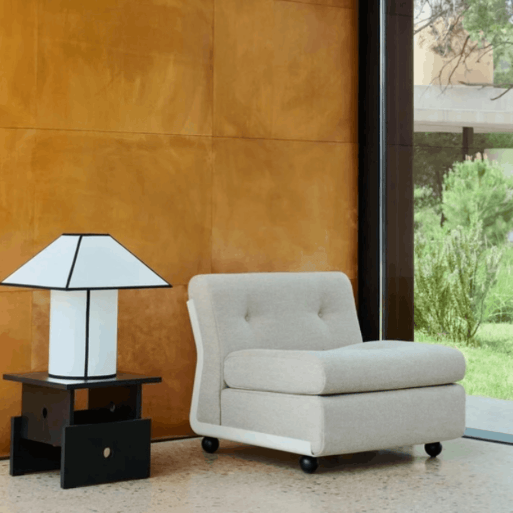 A light beige upholstered armchair with tufted back cushions and black caster wheels, shown in a modern interior next to a black and white table lamp against a warm brown wall.