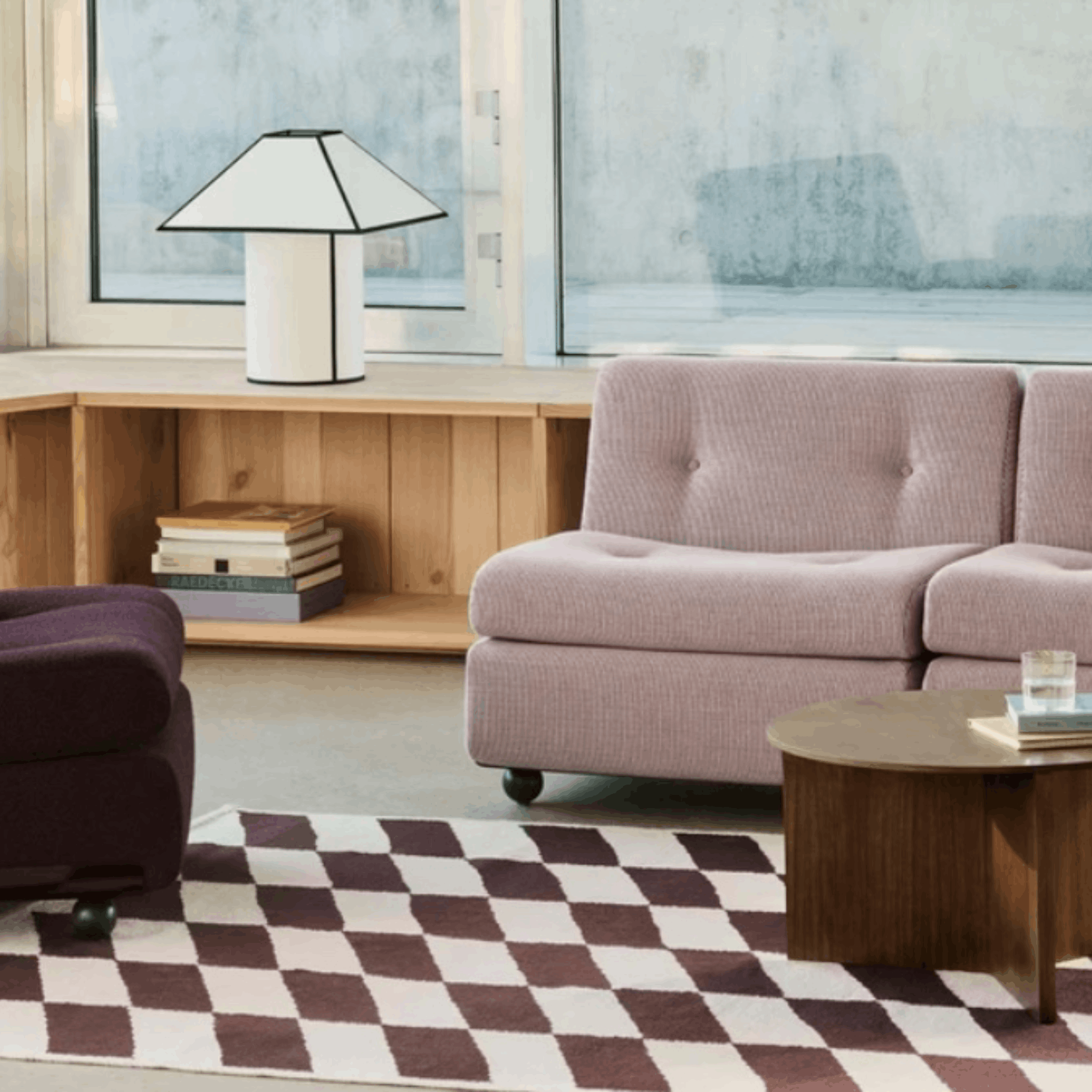 A modern living room scene featuring a light pink tufted sofa in a textured fabric, set next to a dark purple armchair and a checkered rug. A wooden side table and a white table lamp with a black frame are also visible in the room.