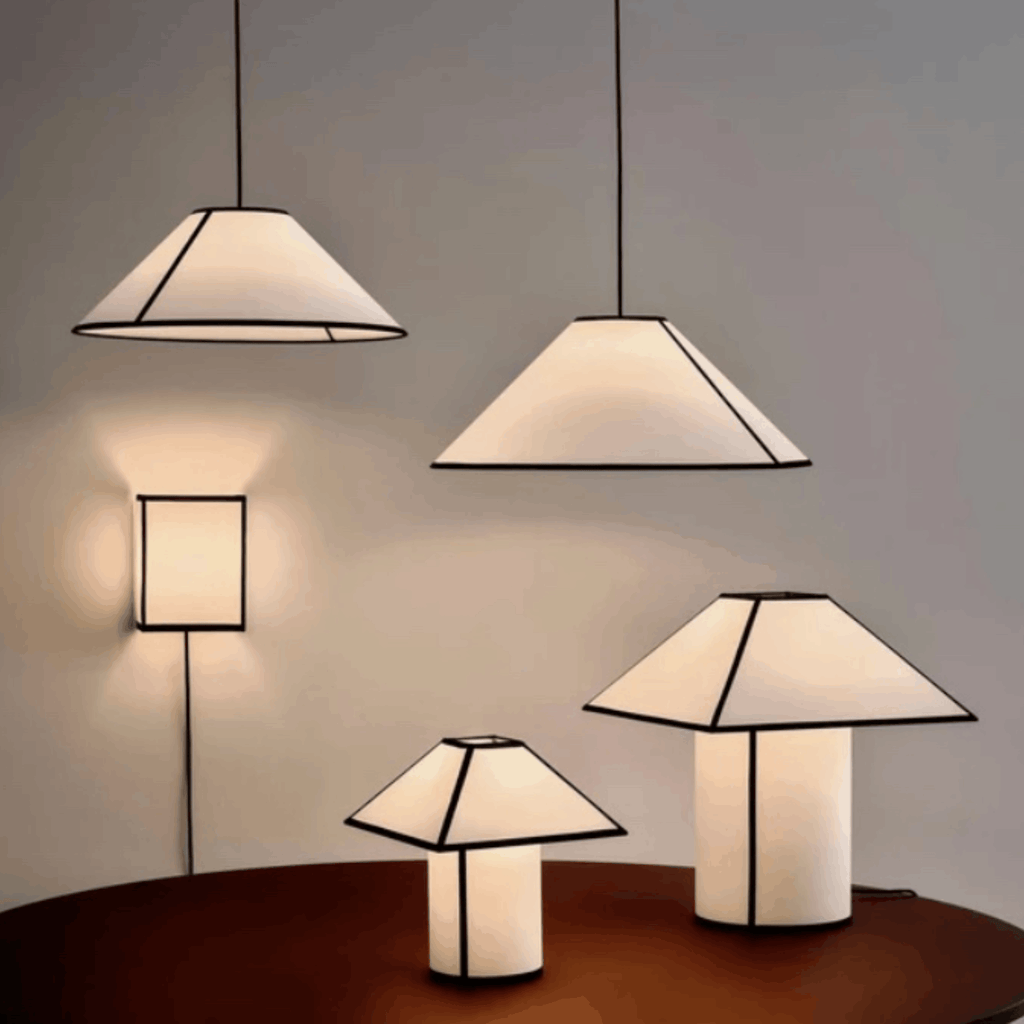 Four modern geometric table and wall lamps with white shades and black metal frames, shown in different sizes against a neutral gray wall and a dark wooden table.