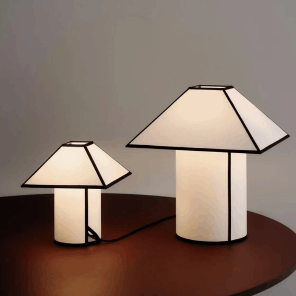 Two modern table lamps with geometric white shades and black metal frames, one small and one large, placed on a dark wooden surface against a neutral gray background.