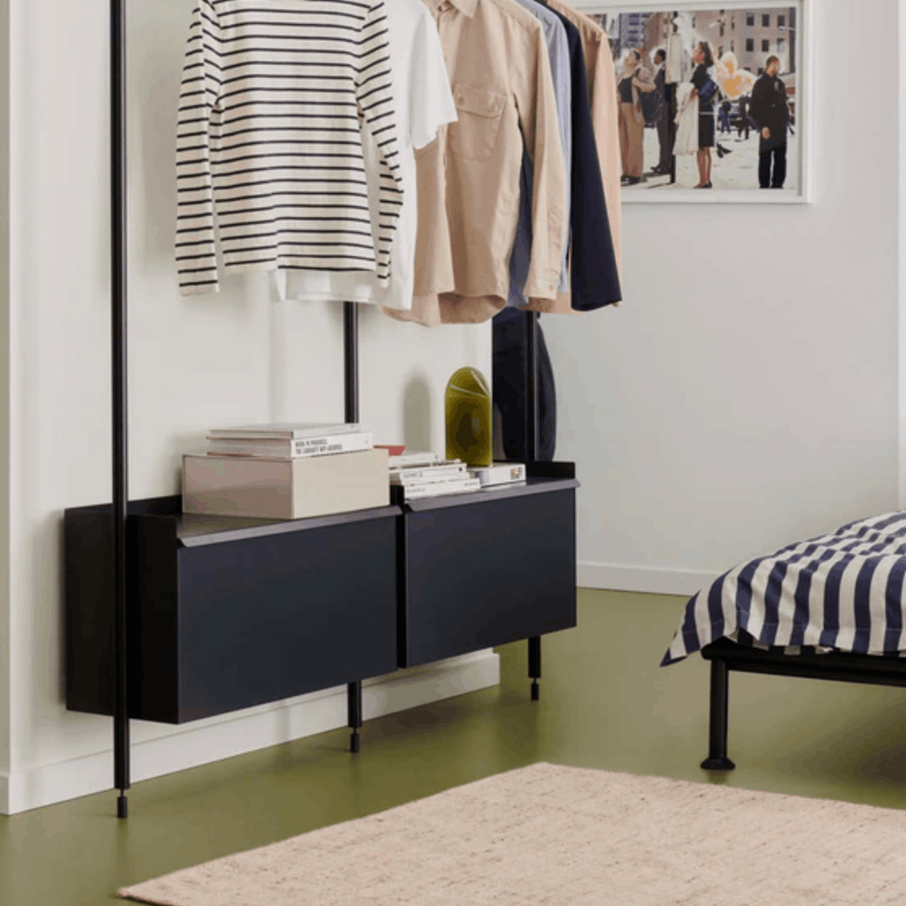 Clothing rack with clothes and a black console table in a room with a bed and picture on the wall. Alongside the bed is a hays crayon effect rug.