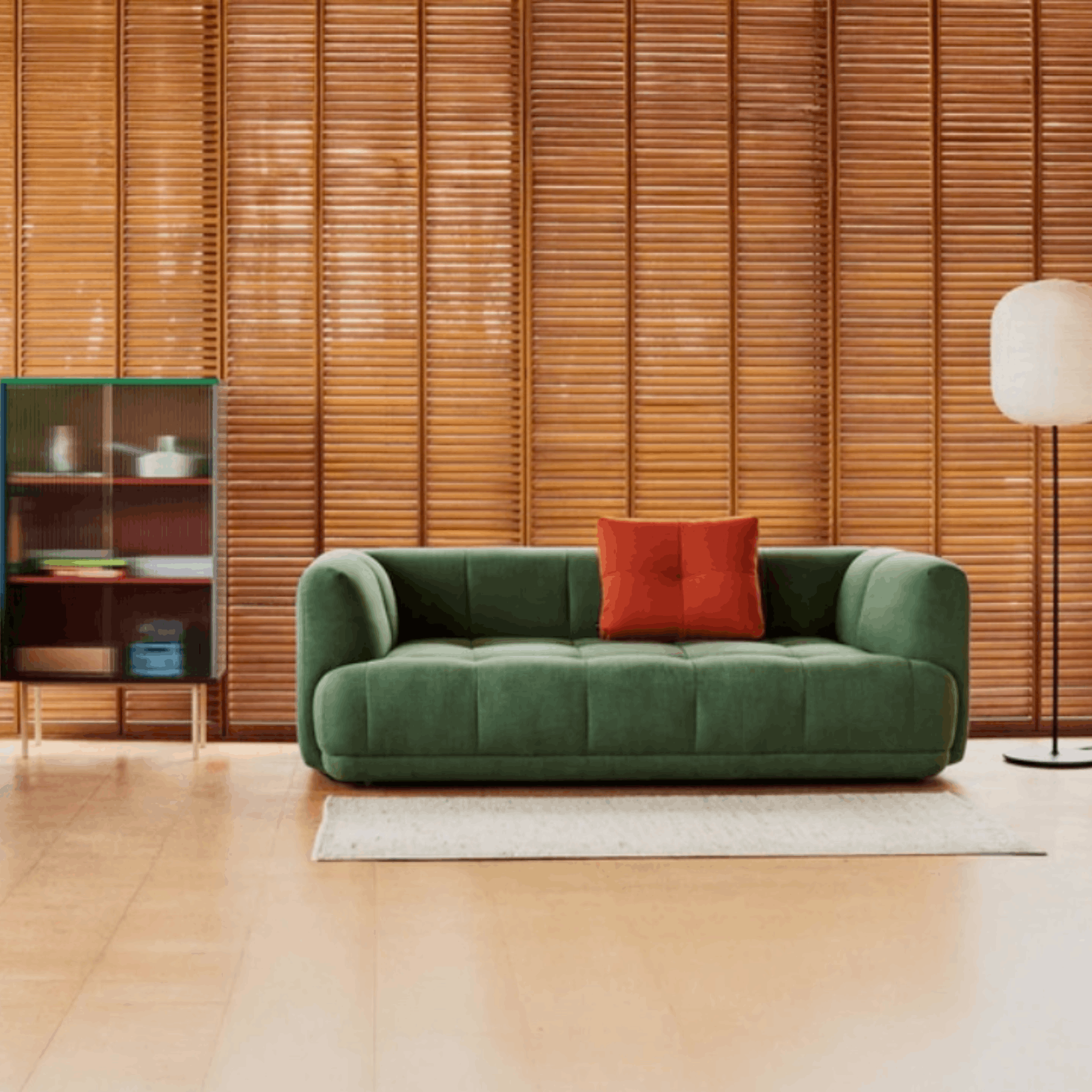 A green tufted velvet loveseat sofa with a red accent pillow, set in a modern living room with a wooden slat wall, light wood floor, a white rug, and a floor lamp.