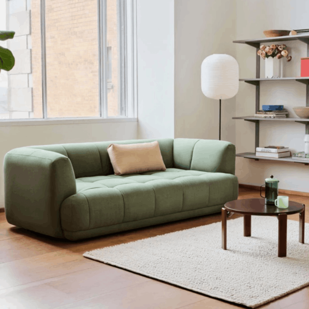 A green tufted loveseat sofa in a bright, modern living room setting with a beige throw pillow, a white shag rug, a small wooden coffee table, and a floor lamp.