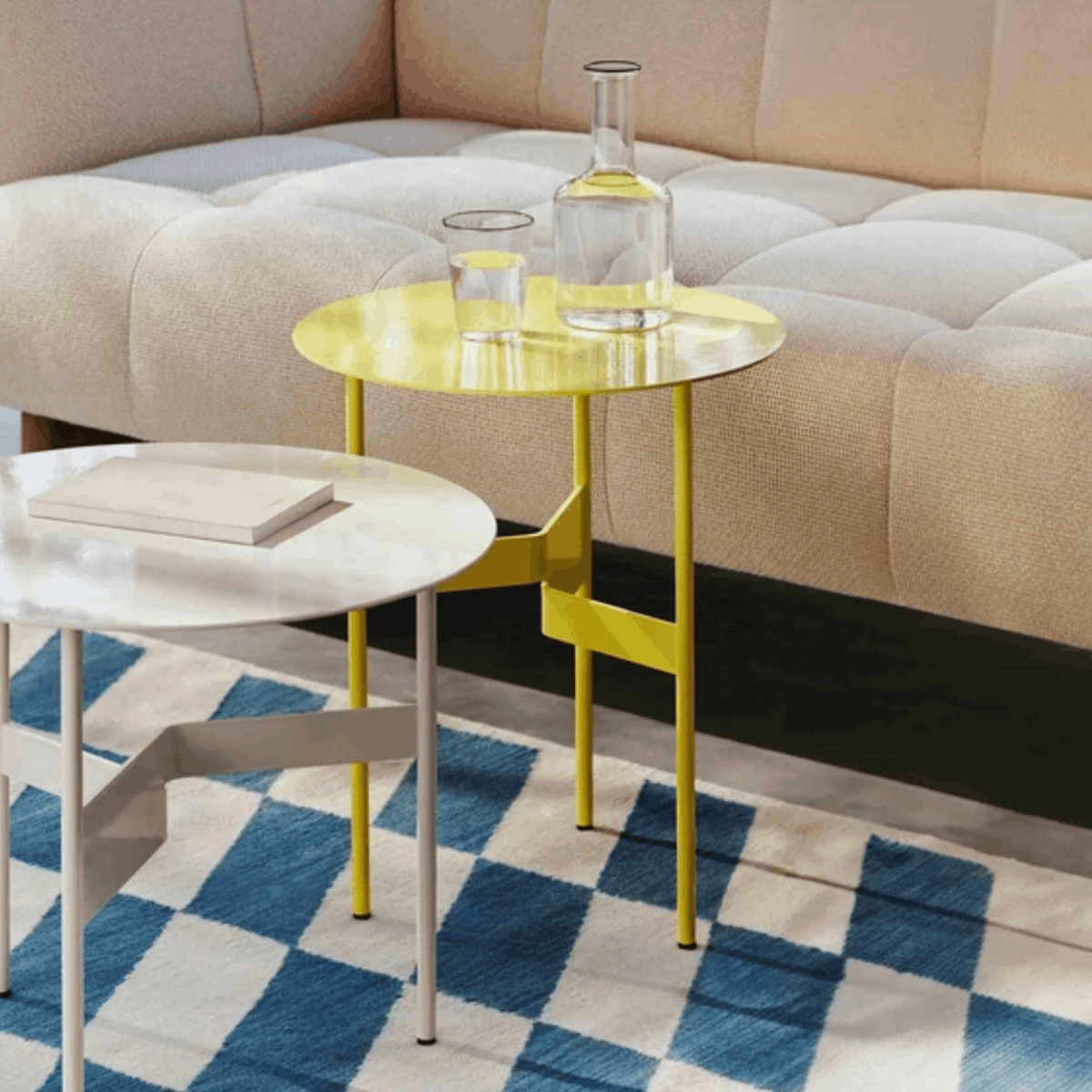 A modern yellow round nesting side table with a glass top and metal legs, styled in a living room next to a beige sofa with a blue and white checkered rug.