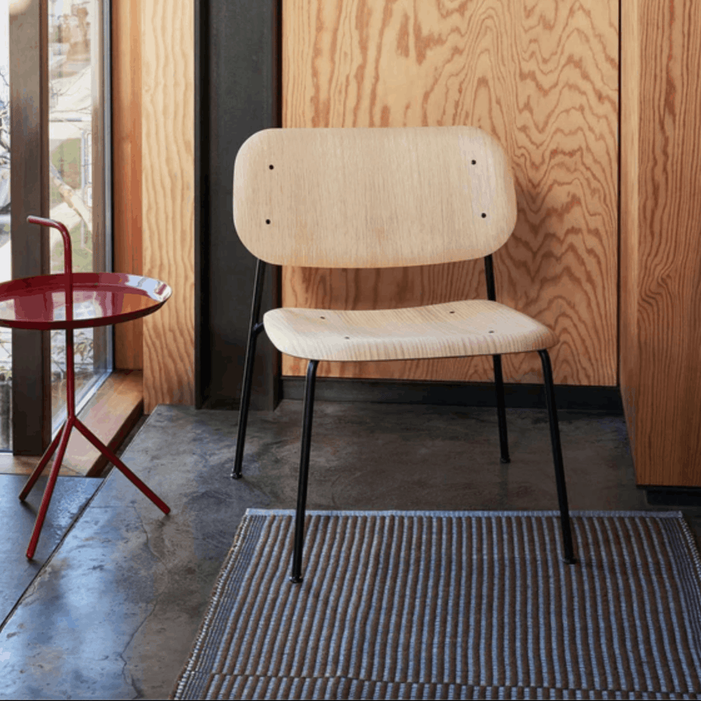 Beige chair with black legs in a room with wooden walls, a red side table and a Hay rug in the floor