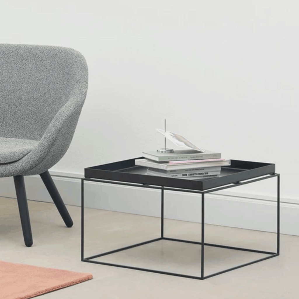 A modern square coffee table with a black metal frame and a dark tray top, styled with stacked books and a small decorative object. The table is shown in a minimalist room setting next to a gray upholstered chair.