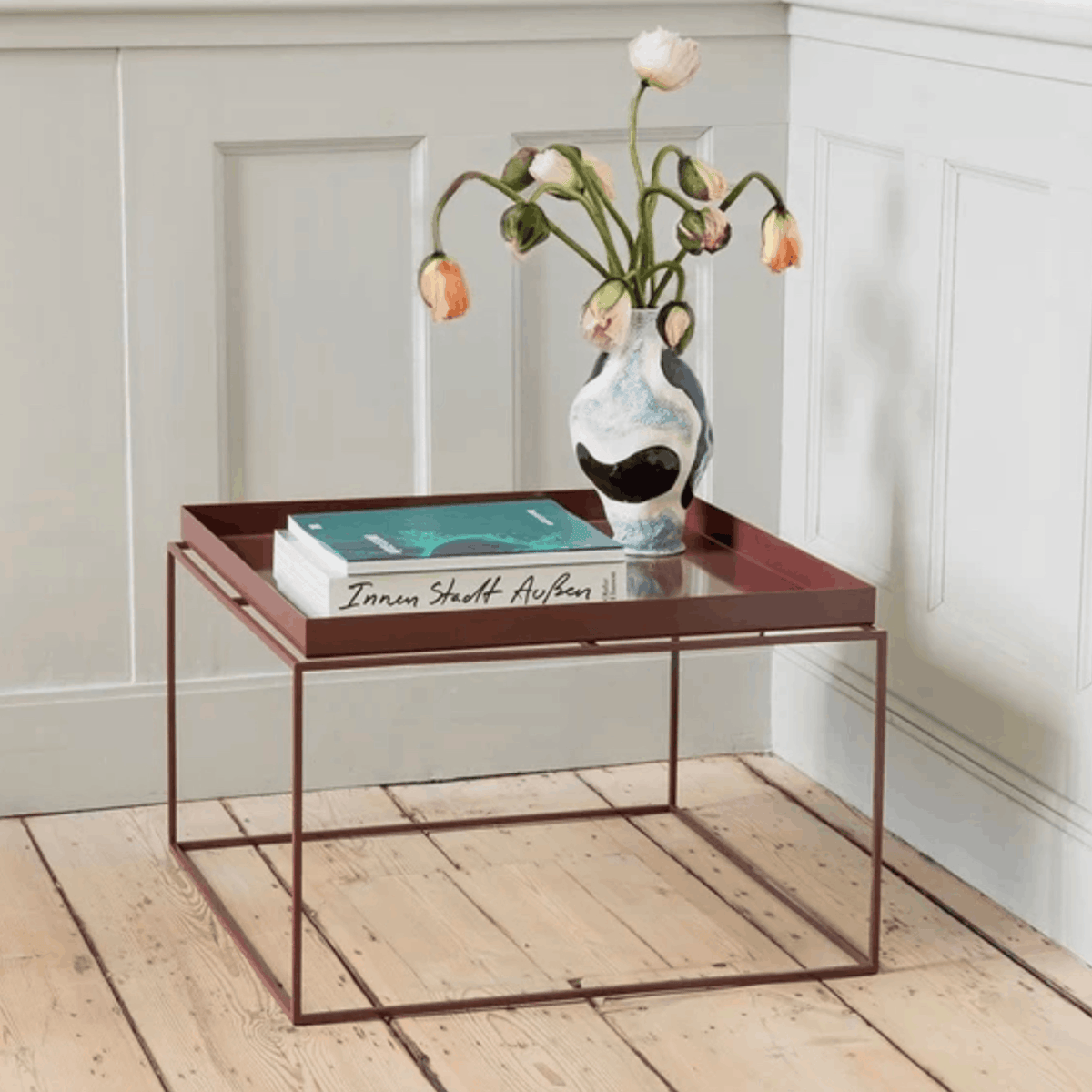 A square metal coffee table with a dark reddish-brown frame and mirrored top, styled with a vase of tulips and books in a bright room with white panelled walls and light wood flooring.