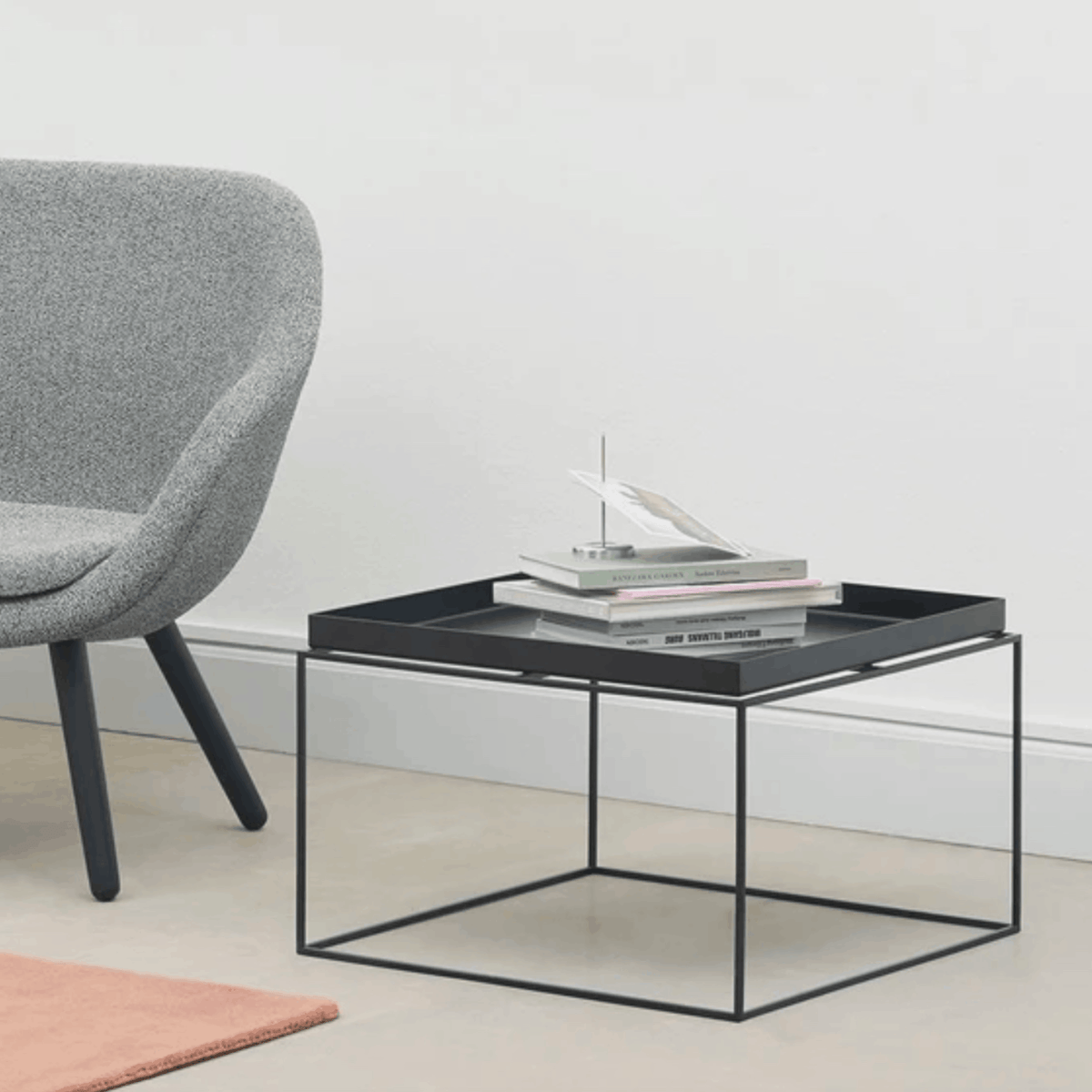 A modern square coffee table with a black metal frame and a dark tray top, styled with stacked books and a small decorative object. The table is shown in a minimalist room setting next to a gray upholstered chair.