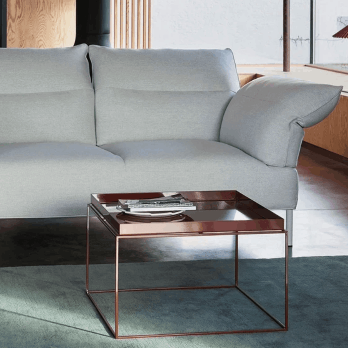 A modern square coffee table with a copper-toned metal frame and a dark glass top, styled in a living room next to a light gray sofa.