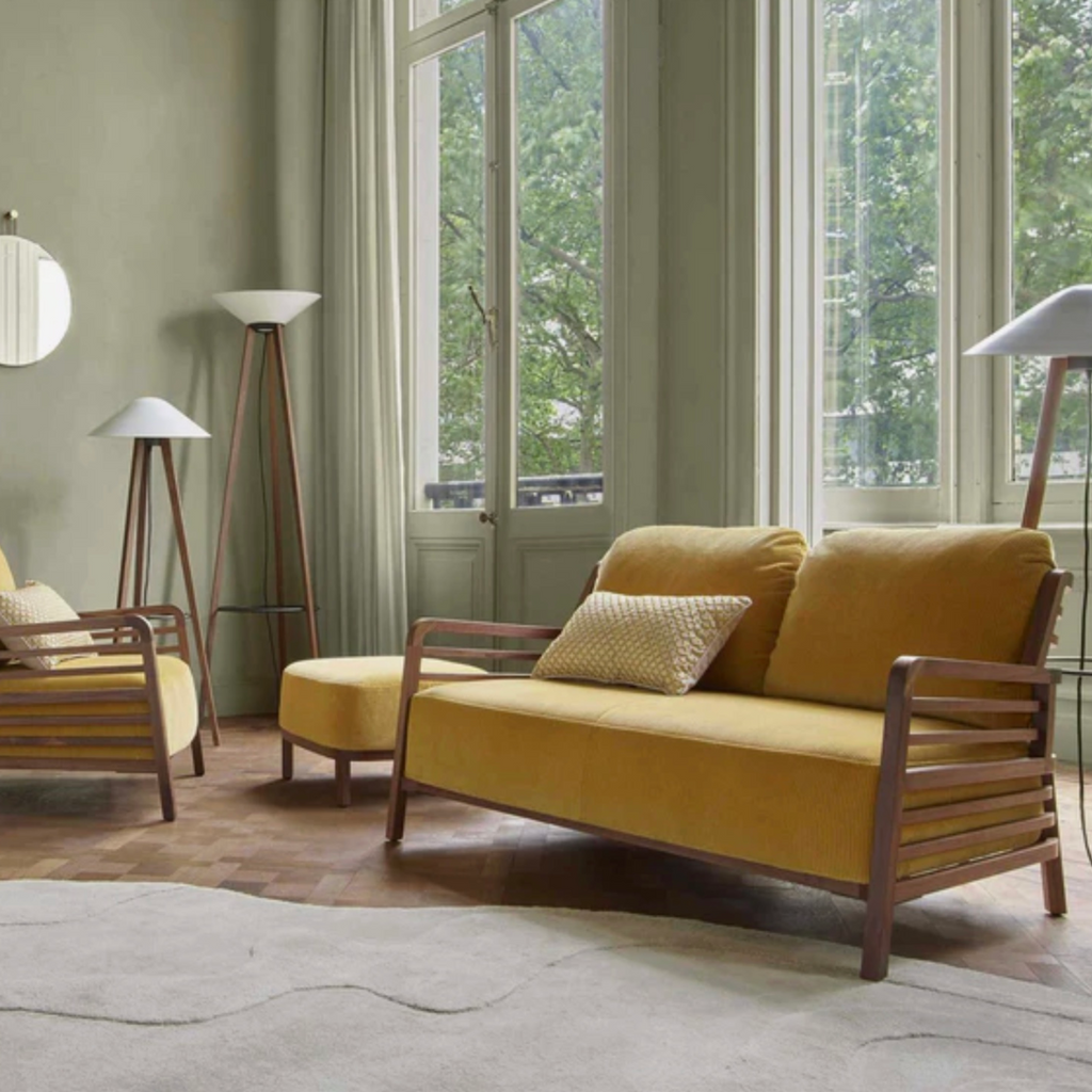Yellow sofa with wooden frame on a Ligne Roset Gavrinis beige rug in a room with large windows and greenery outside