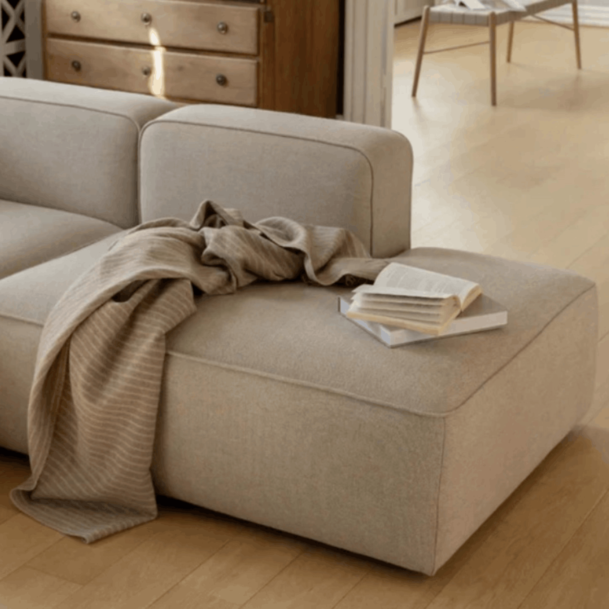 A modern modular sectional sofa in a light beige linen fabric, styled with a draped beige knit throw blanket and a stack of books on the chaise lounge, set in a warm, sunlit room with light wood floors.